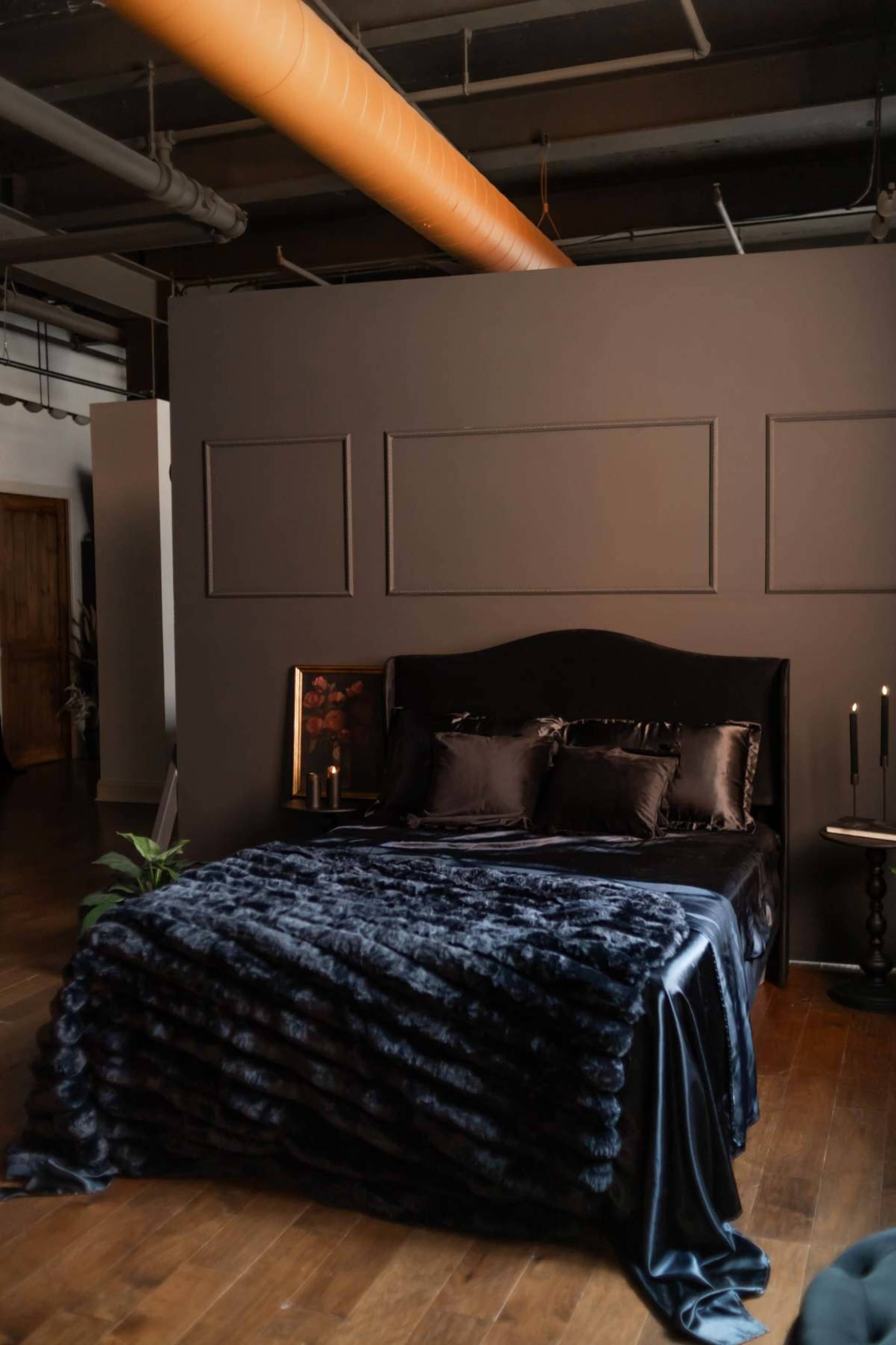 The image shows a dark-themed bedroom with a large, plush bed covered in a textured blanket, accompanied by decorative pillows and a bedside table with candles.