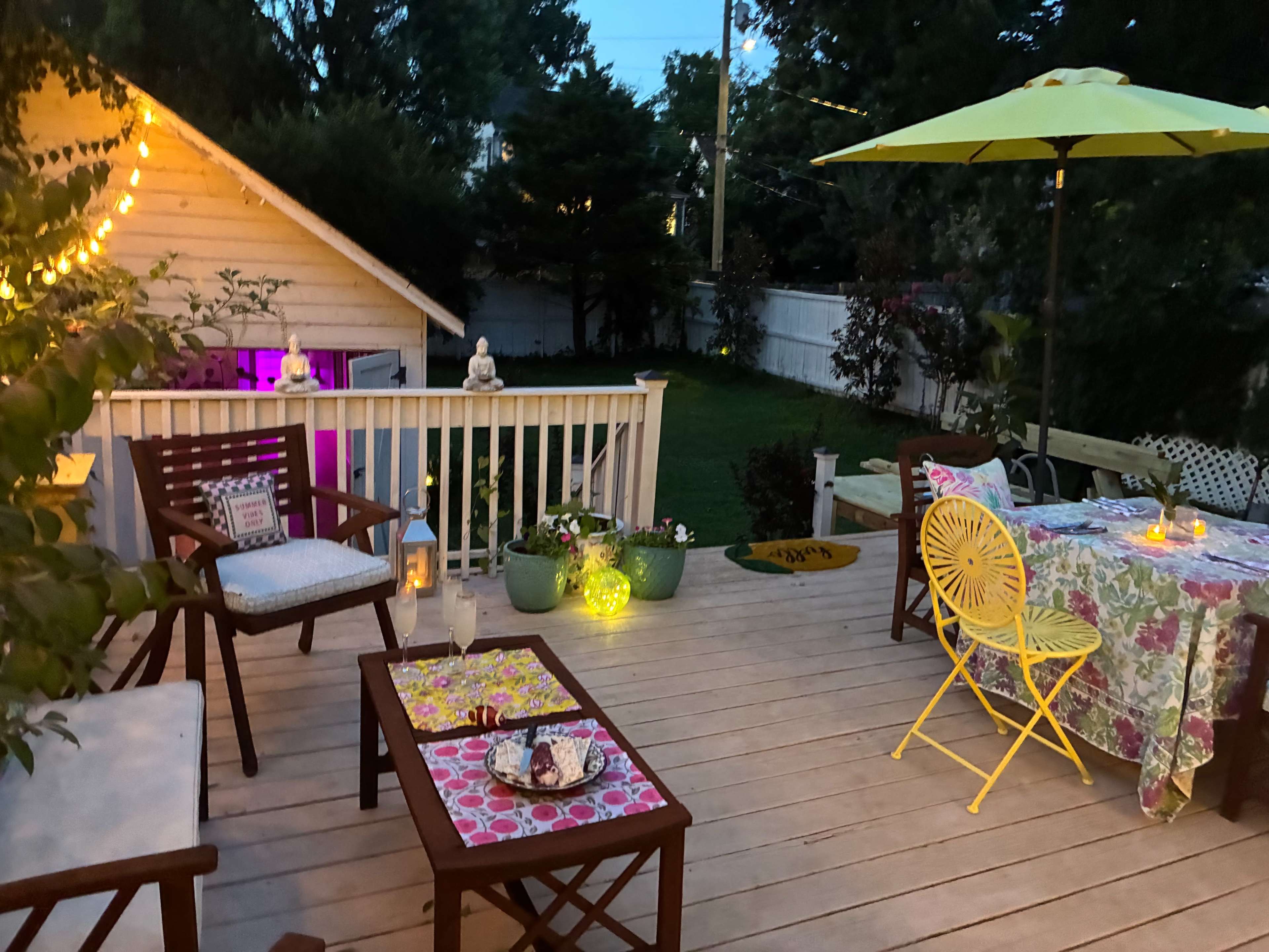 A well-decorated outdoor deck features colorful furniture, plants, and ambient lighting, creating a cozy seating area near a garden.