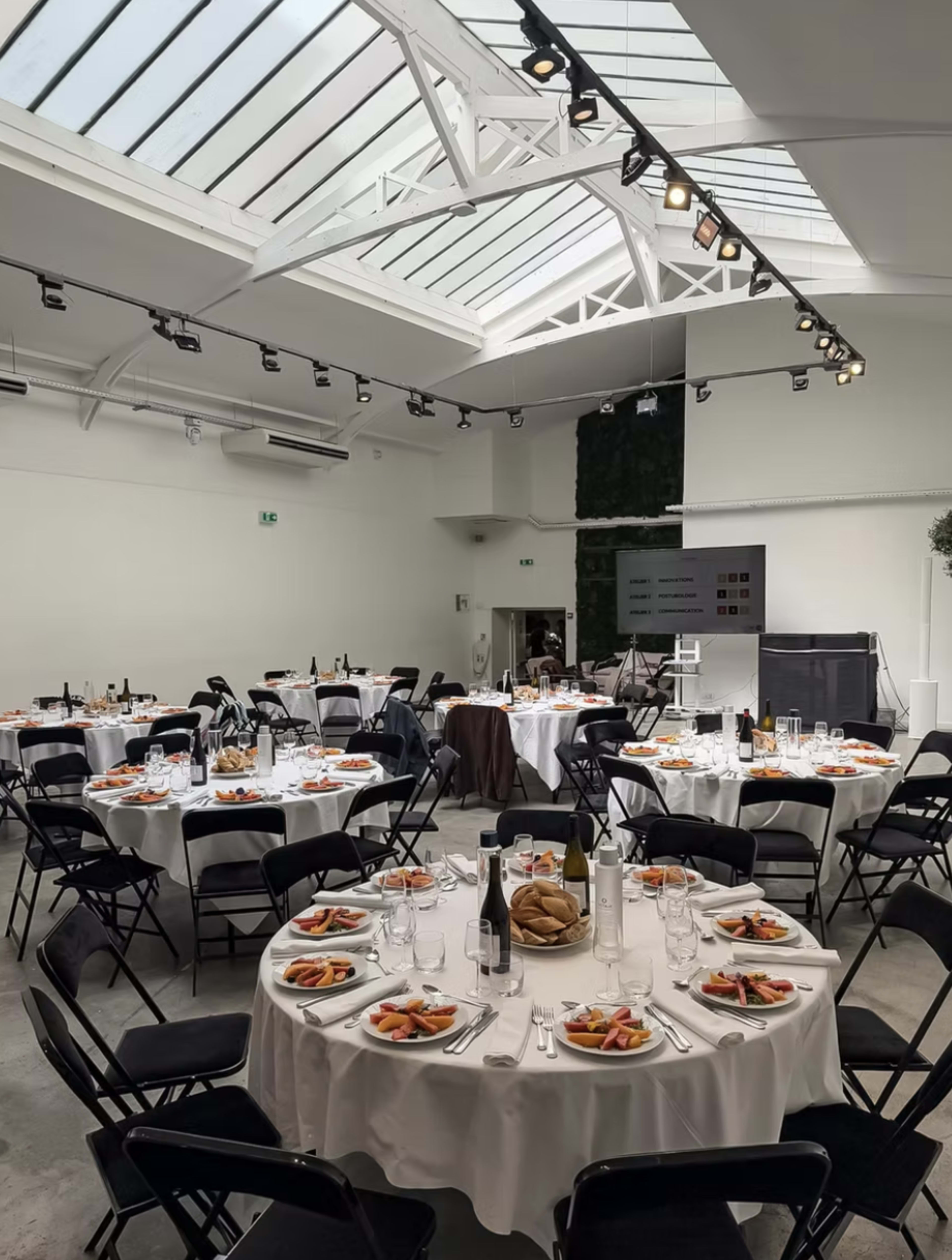 A spacious event room features multiple round tables set with dishes and glasses, surrounded by black chairs and illuminated by natural light from a skylight.