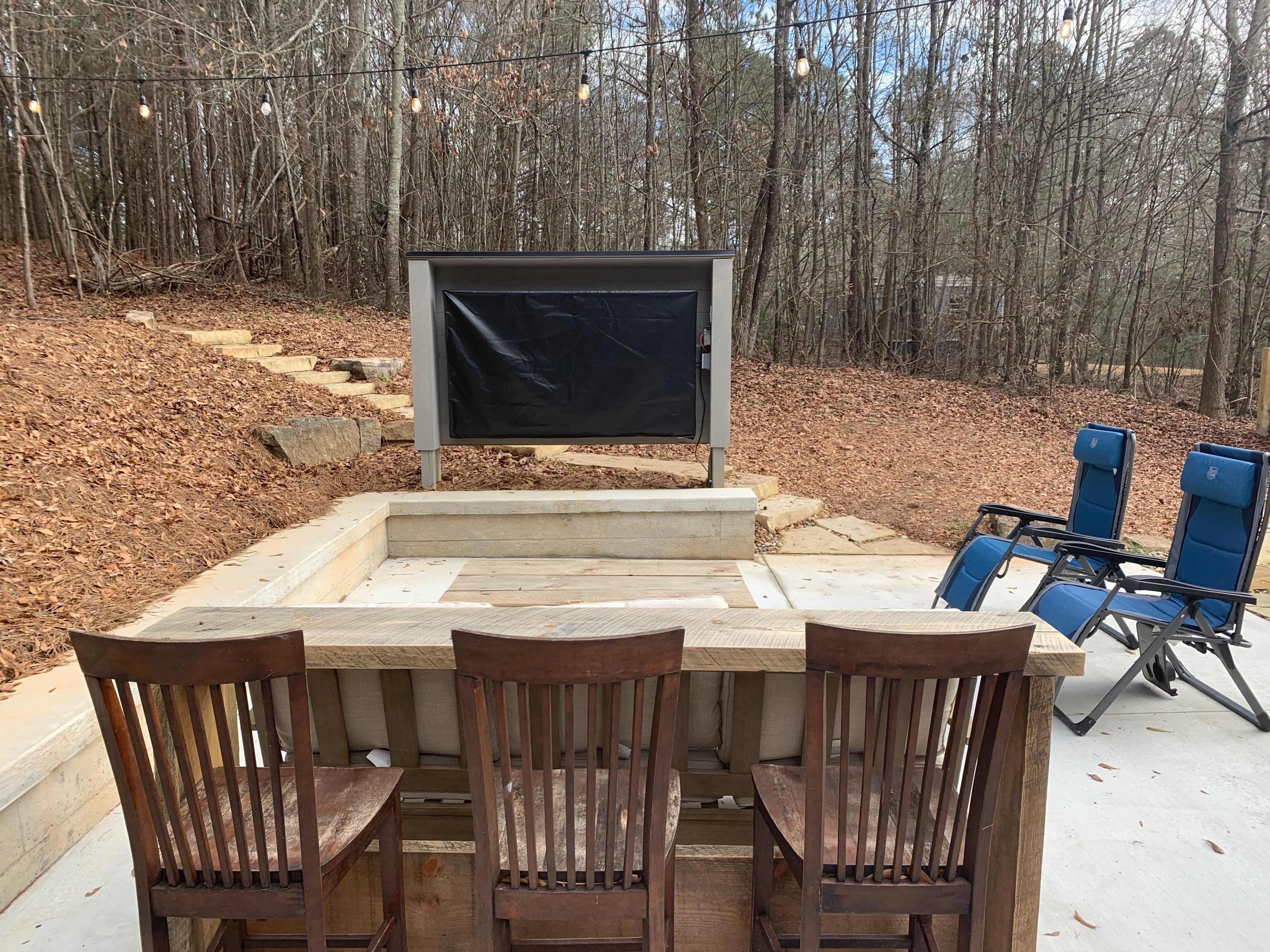 The image shows a backyard seating area with four wooden chairs facing a raised platform that has a black screen, surrounded by trees with fallen leaves on the ground.