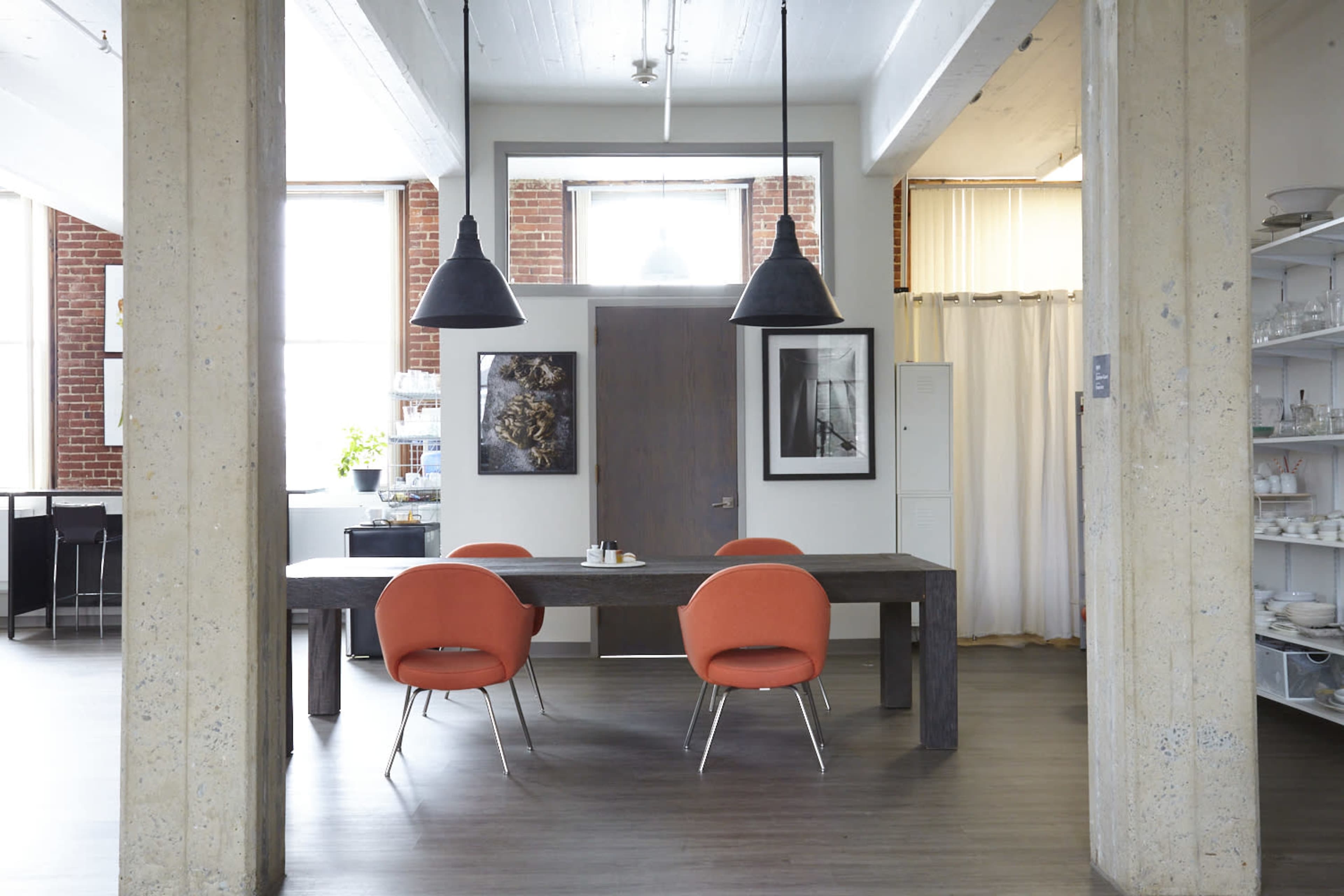 A modern dining area featuring a large wooden table with four orange chairs, two pendant lights hanging from the ceiling, and large windows allowing natural light to enter.
