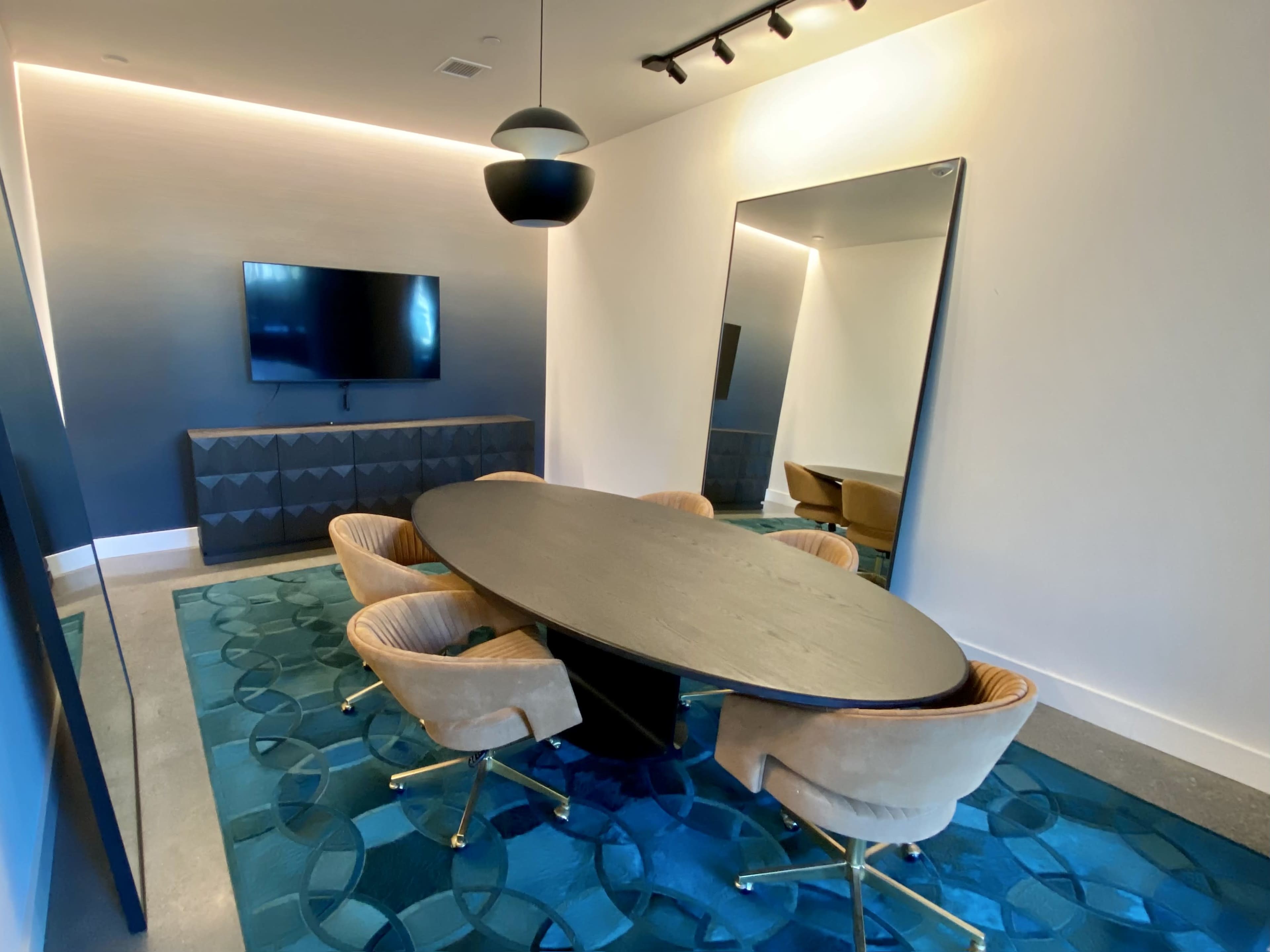 A modern conference room features an oval table surrounded by four upholstered chairs, a wall-mounted television, and a large mirror reflecting the space.