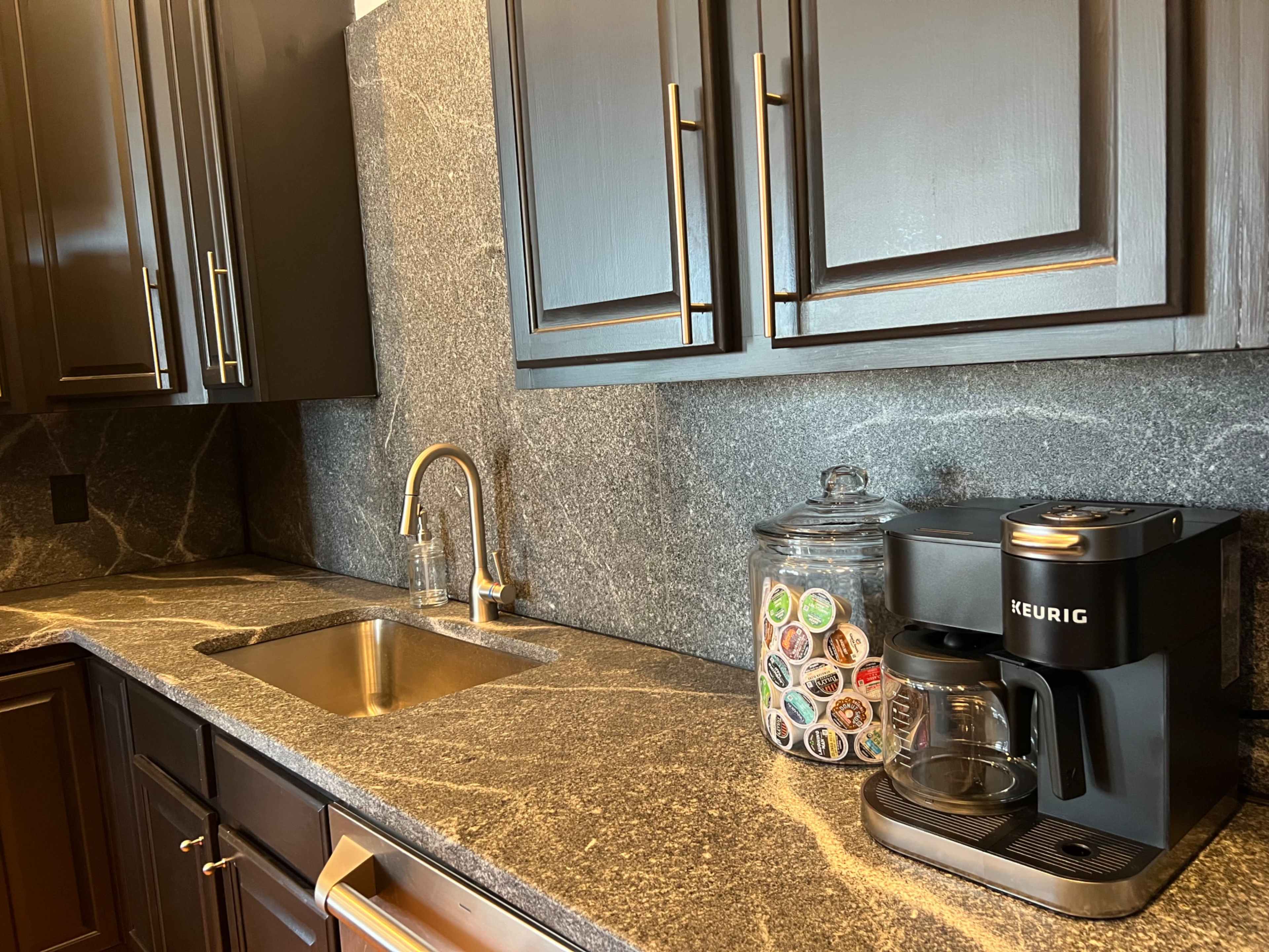 The image shows a modern kitchen countertop featuring a sink, a Keurig coffee maker, and a jar filled with coffee pods against a dark stone backdrop.