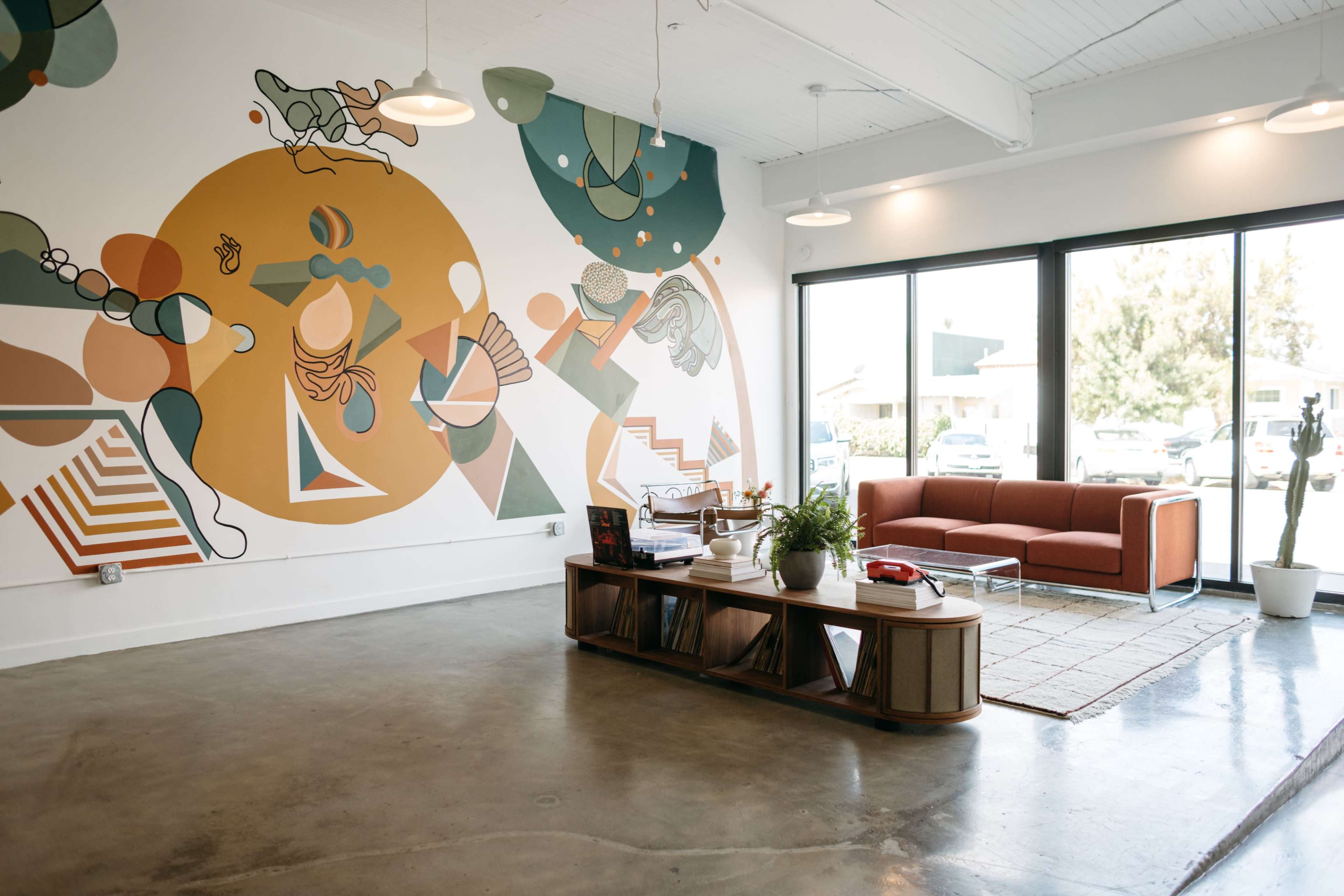 The scene features a modern lounge area with a large abstract mural on the wall, a brown sofa, a low coffee table, and large windows allowing natural light to filter in.