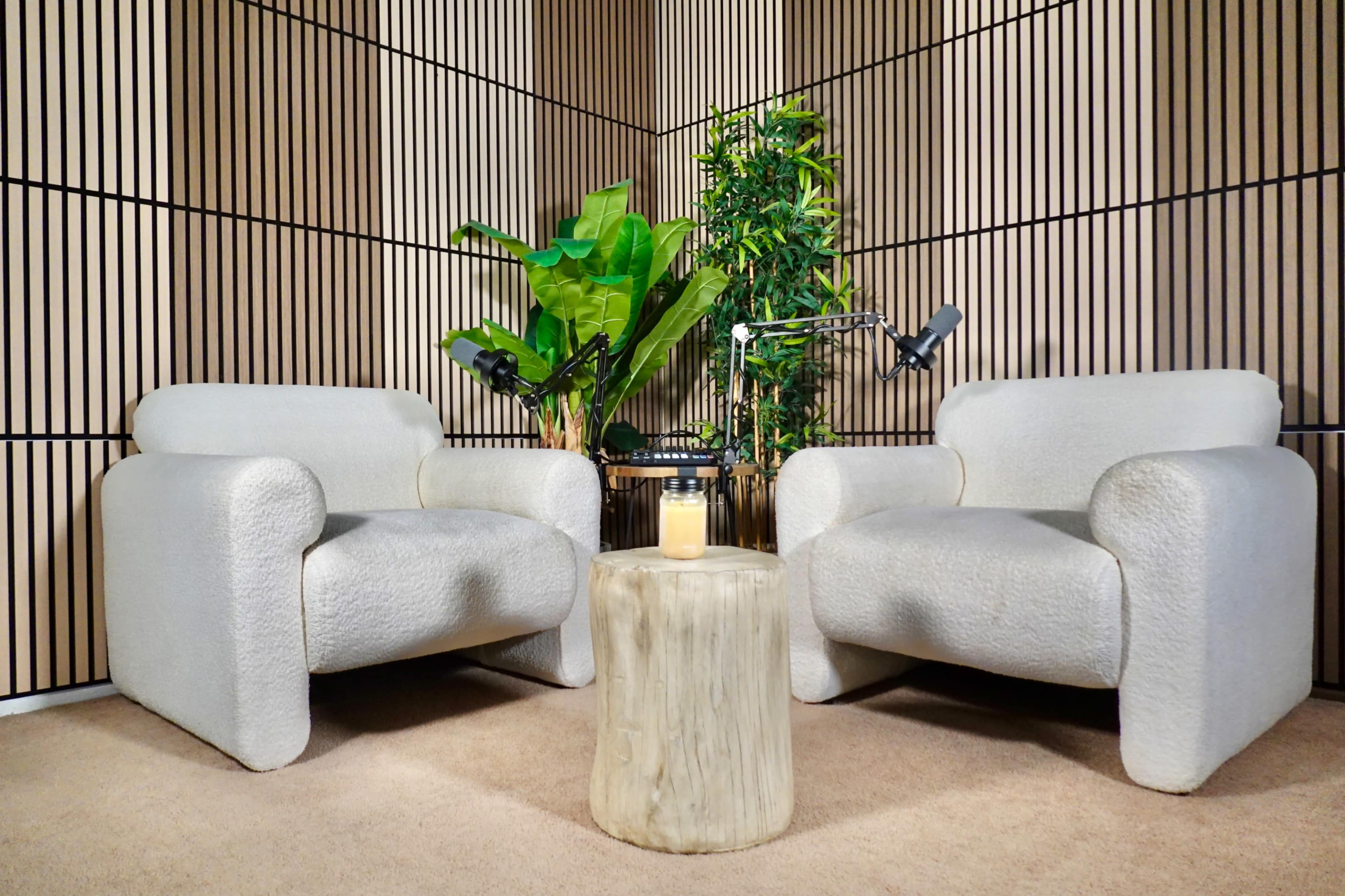 The image shows a modern seating area with two light-colored armchairs, a small round wooden table in the center, and greenery in the background against a striped wooden wall.