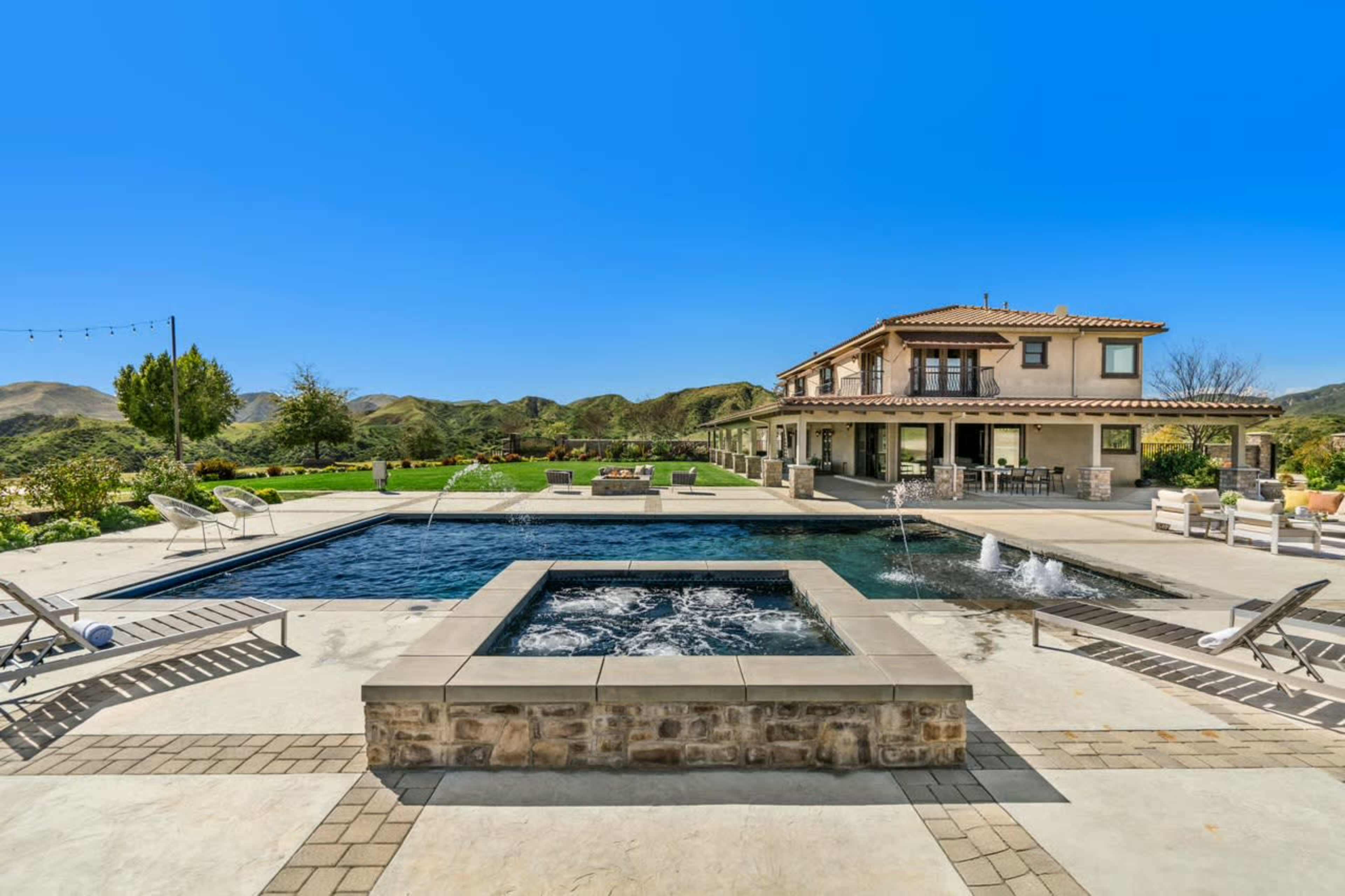 The image shows a modern house with a large swimming pool and spa, set against a backdrop of rolling hills under a clear blue sky.