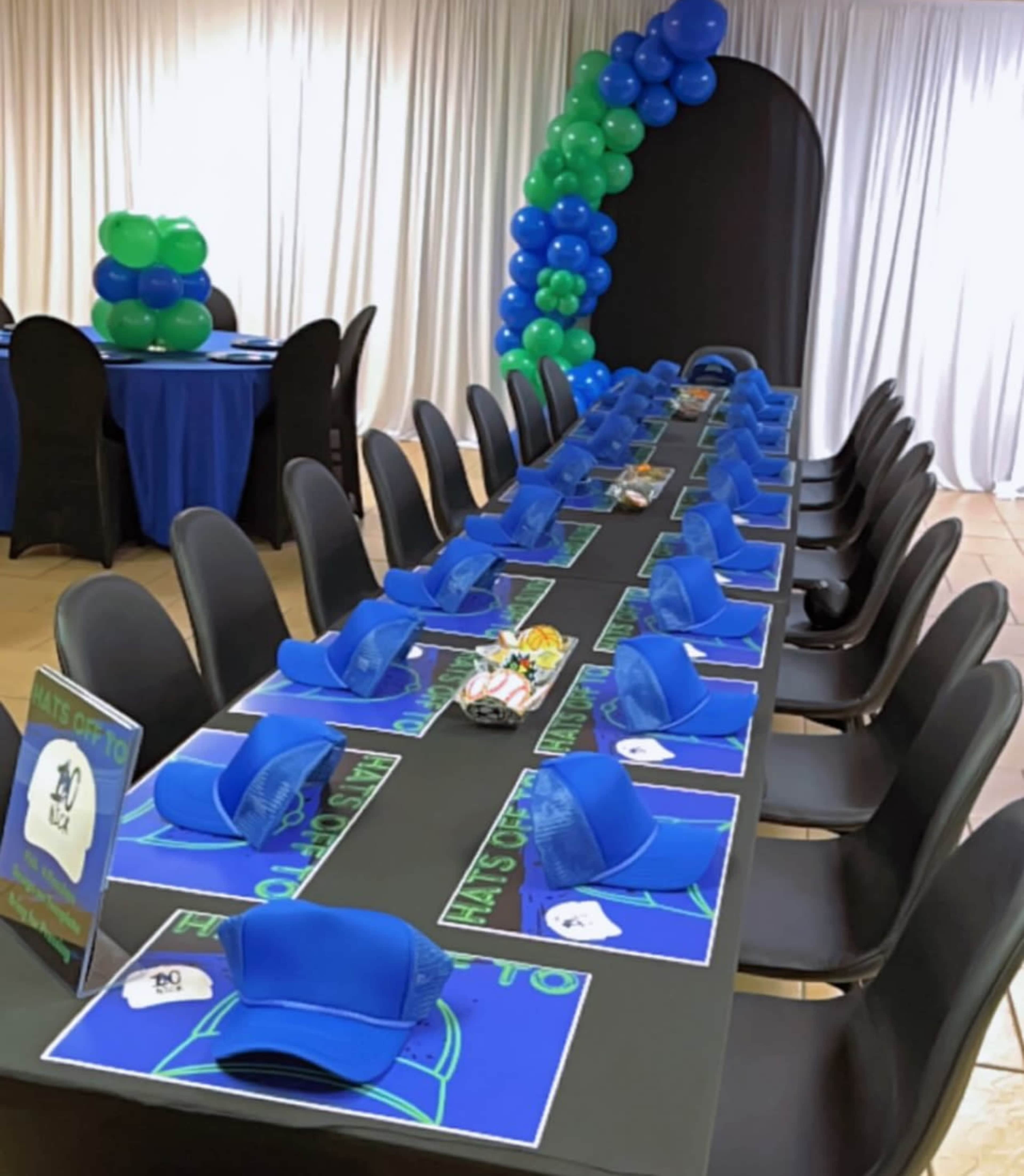 A long rectangular table is set with blue tablecloths, blue hats for each seat, and a colorful balloon arch in the background.
