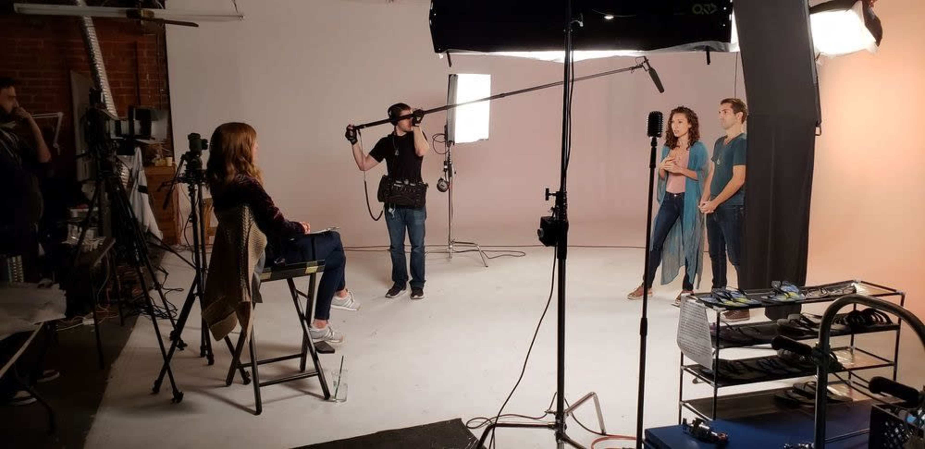 A film crew sets up in a studio with lights and cameras while a male and female actor prepare to perform.