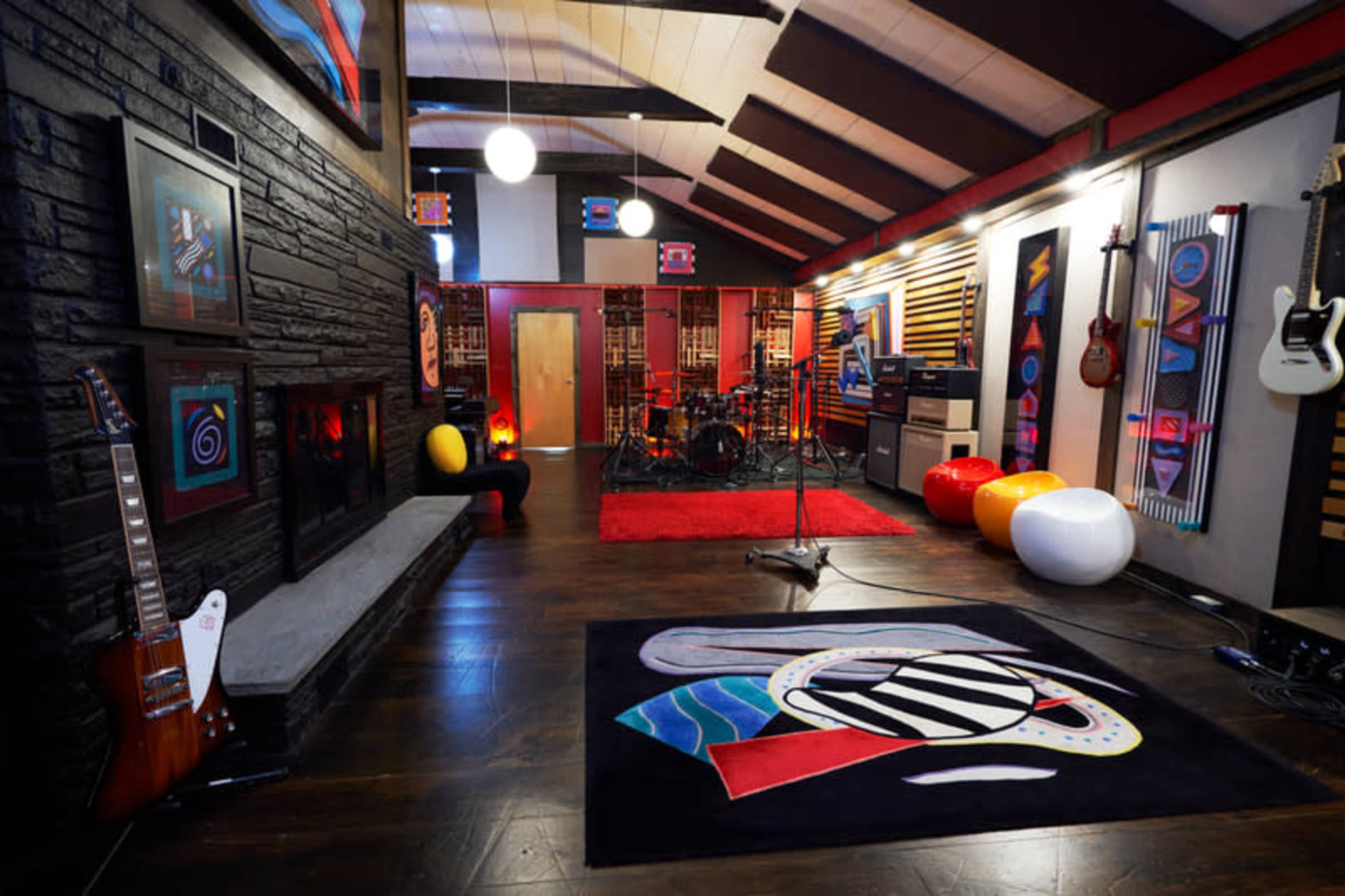 A music studio features a drum set, colorful guitars, and vibrant artwork on the walls, with a plush rug and modern lighting.