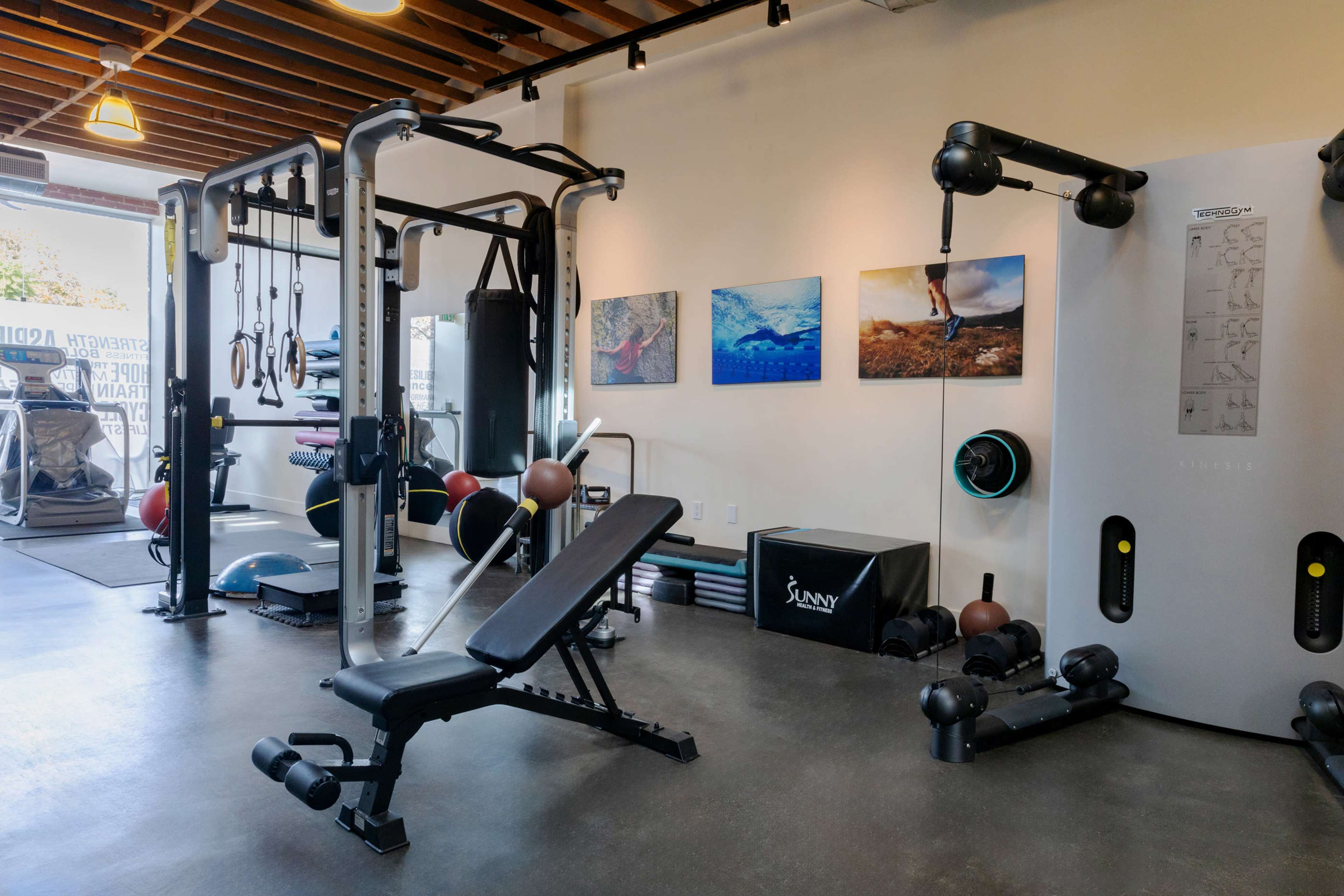 A modern gym interior featuring various fitness equipment, including a multi-station workout machine, a weight bench, and stability balls, with framed photographs on the walls.
