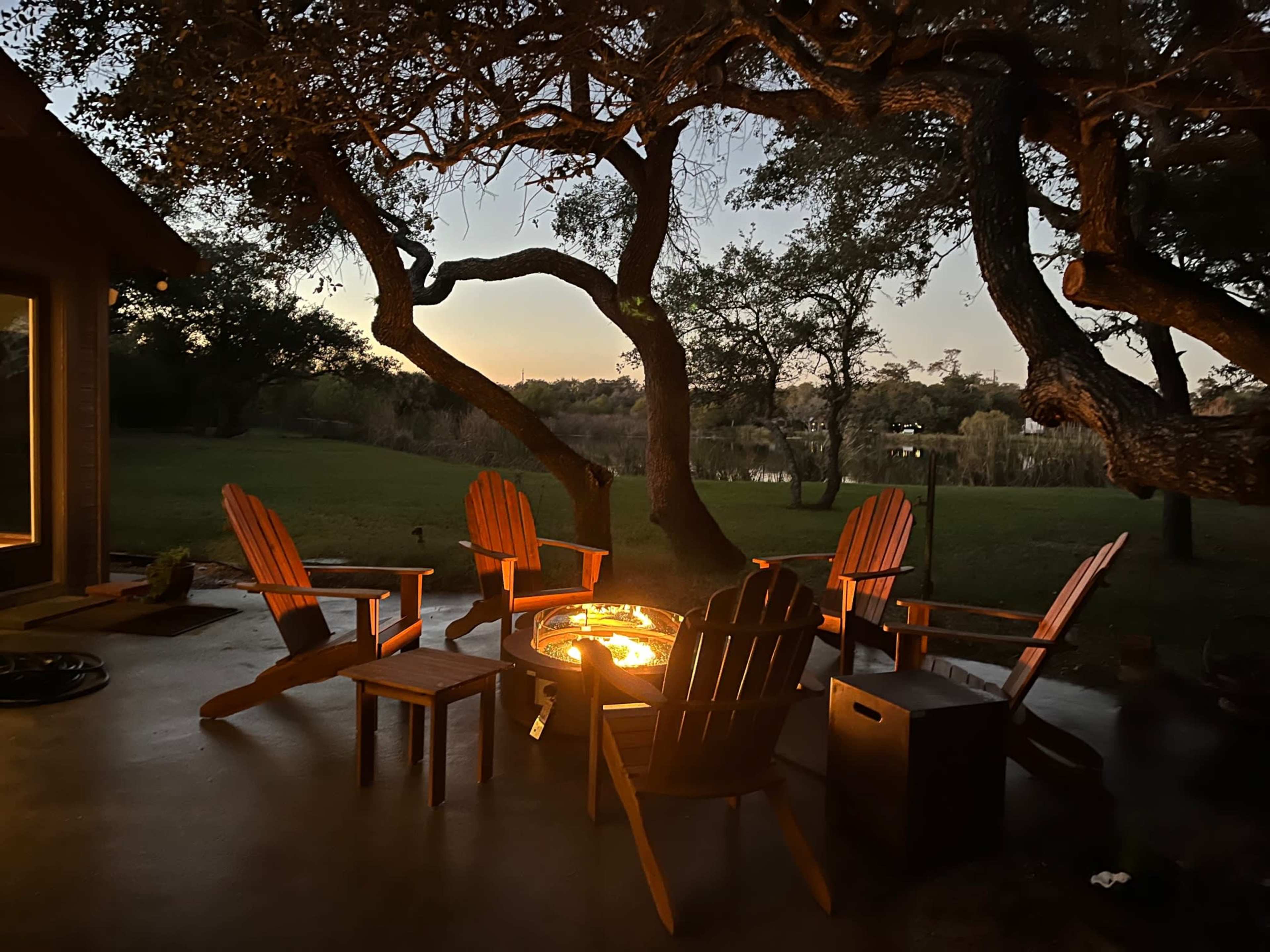 A circle of wooden chairs surrounds a fire pit under large trees at dusk, overlooking a grassy area and water in the background.