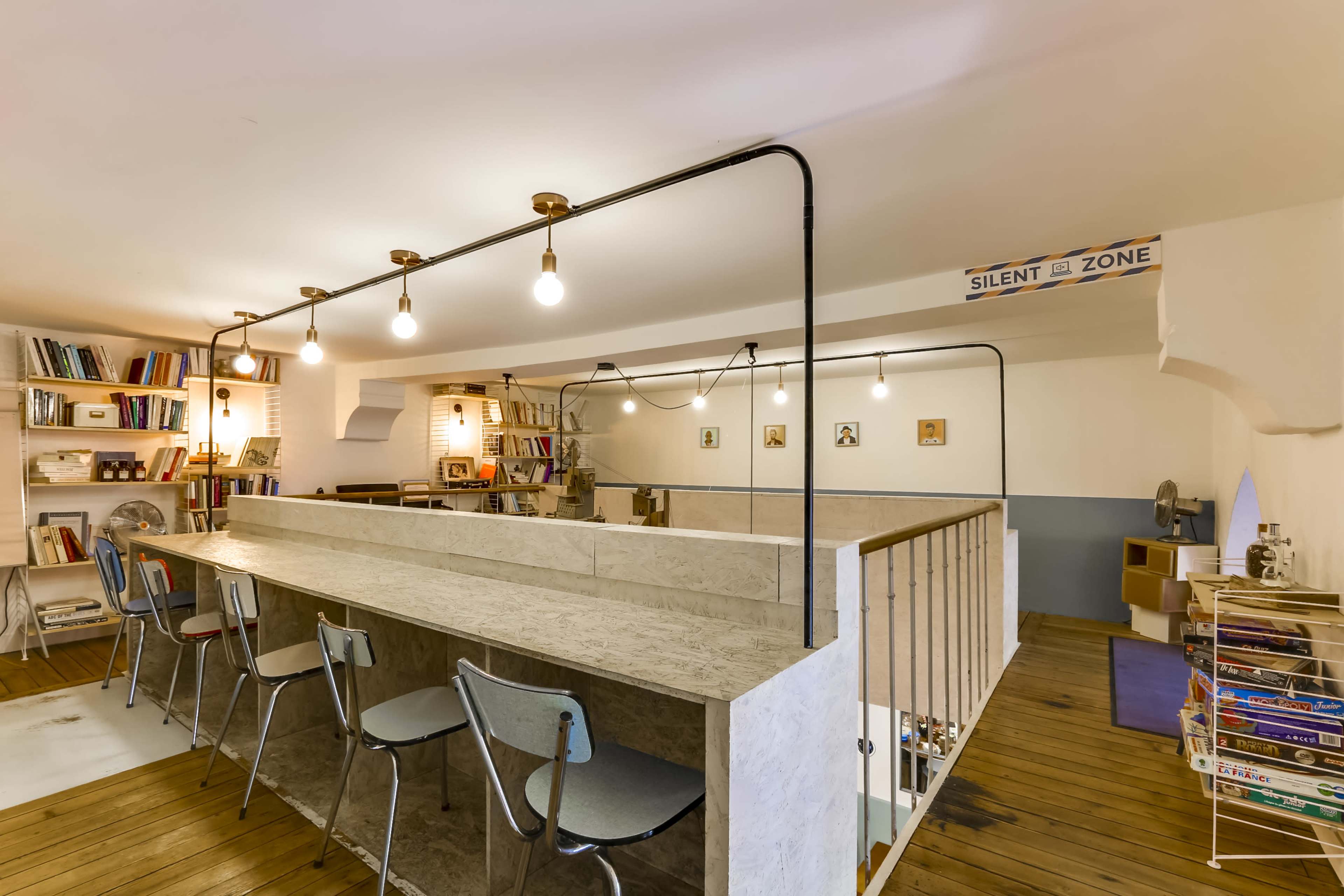 A brightly lit interior space featuring a long, minimalist bar with hanging light bulbs, surrounded by shelves of books and seating.