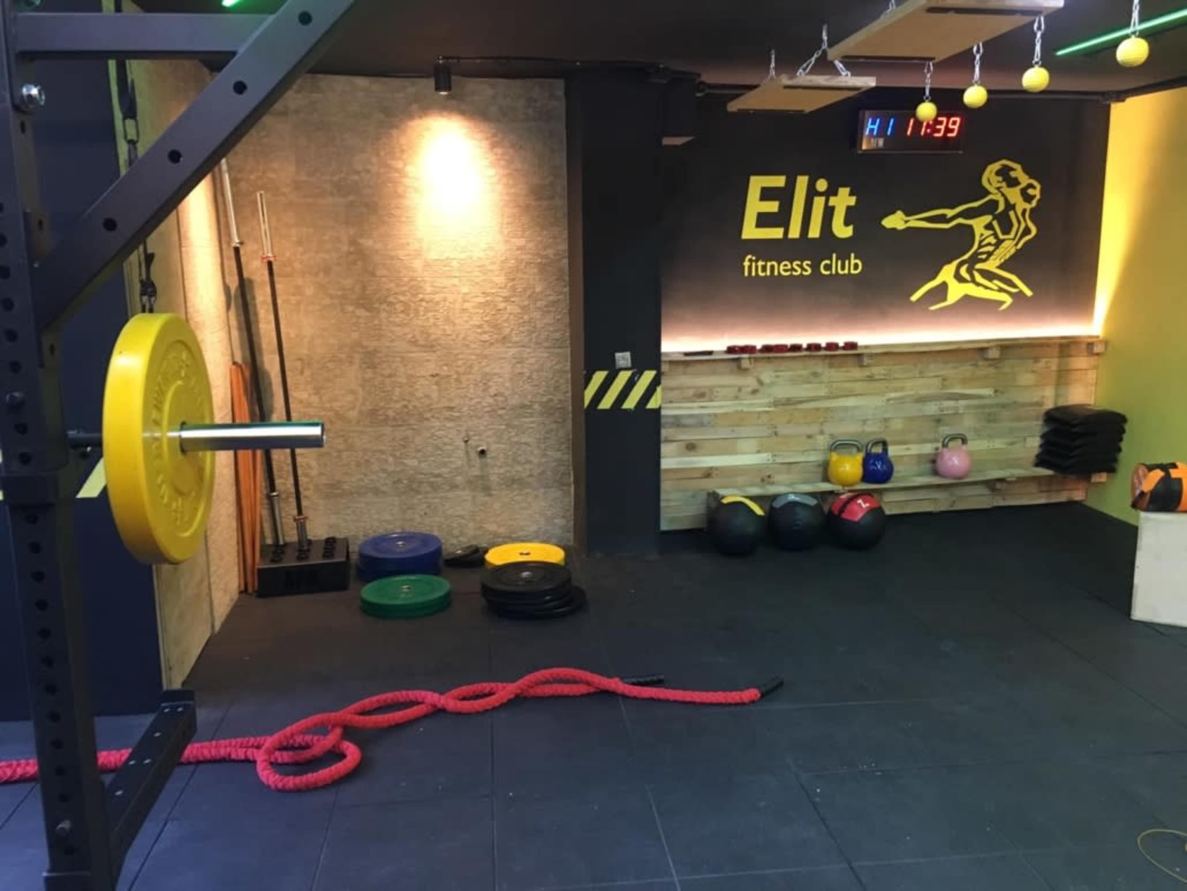 The interior of a fitness club featuring various weights, kettlebells, and exercise ropes situated in a well-lit space with a wall sign that reads "Elit fitness club."