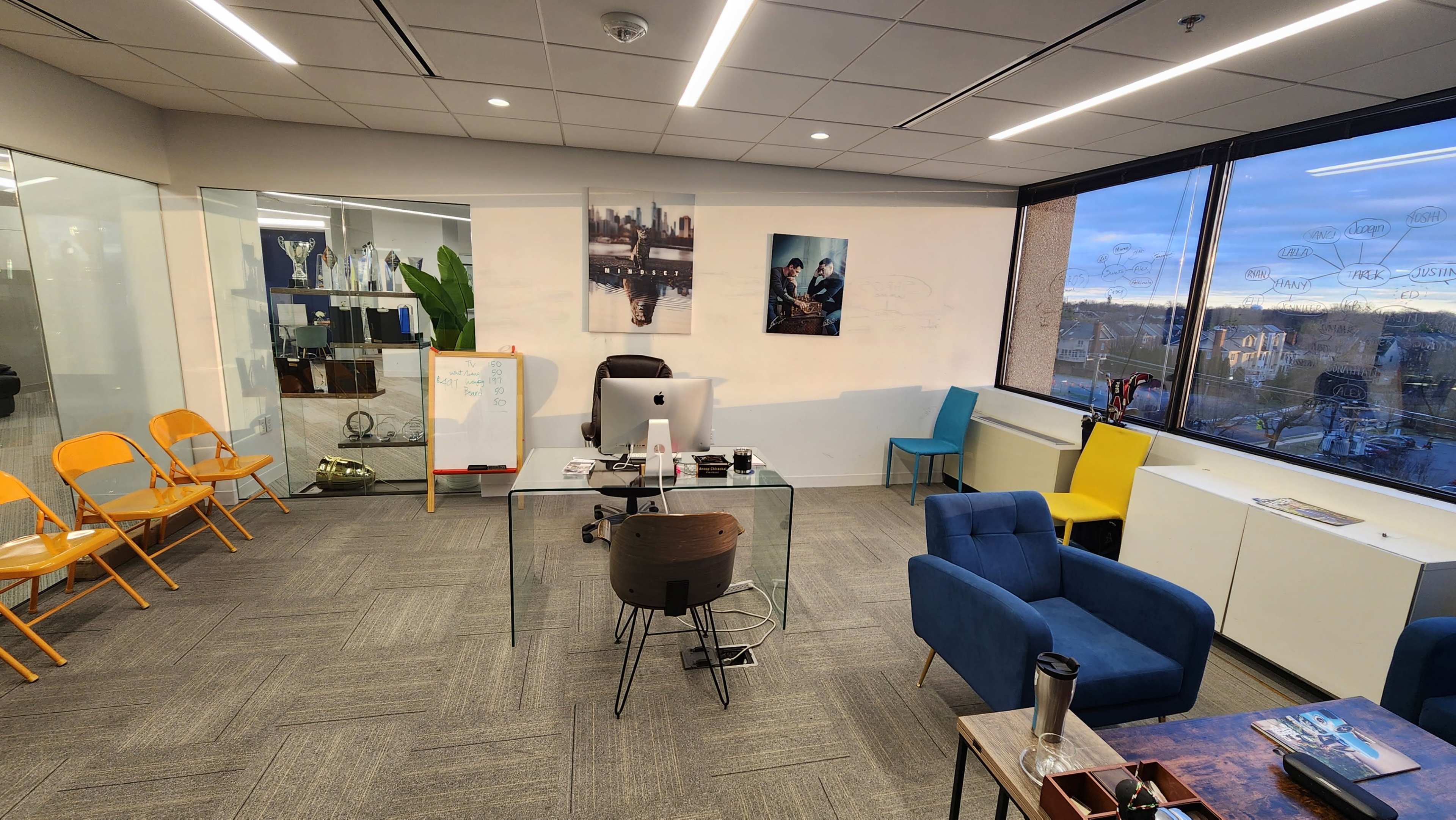 A modern office space with a clear glass desk, colorful chairs, a computer setup, and large windows revealing an outdoor view.