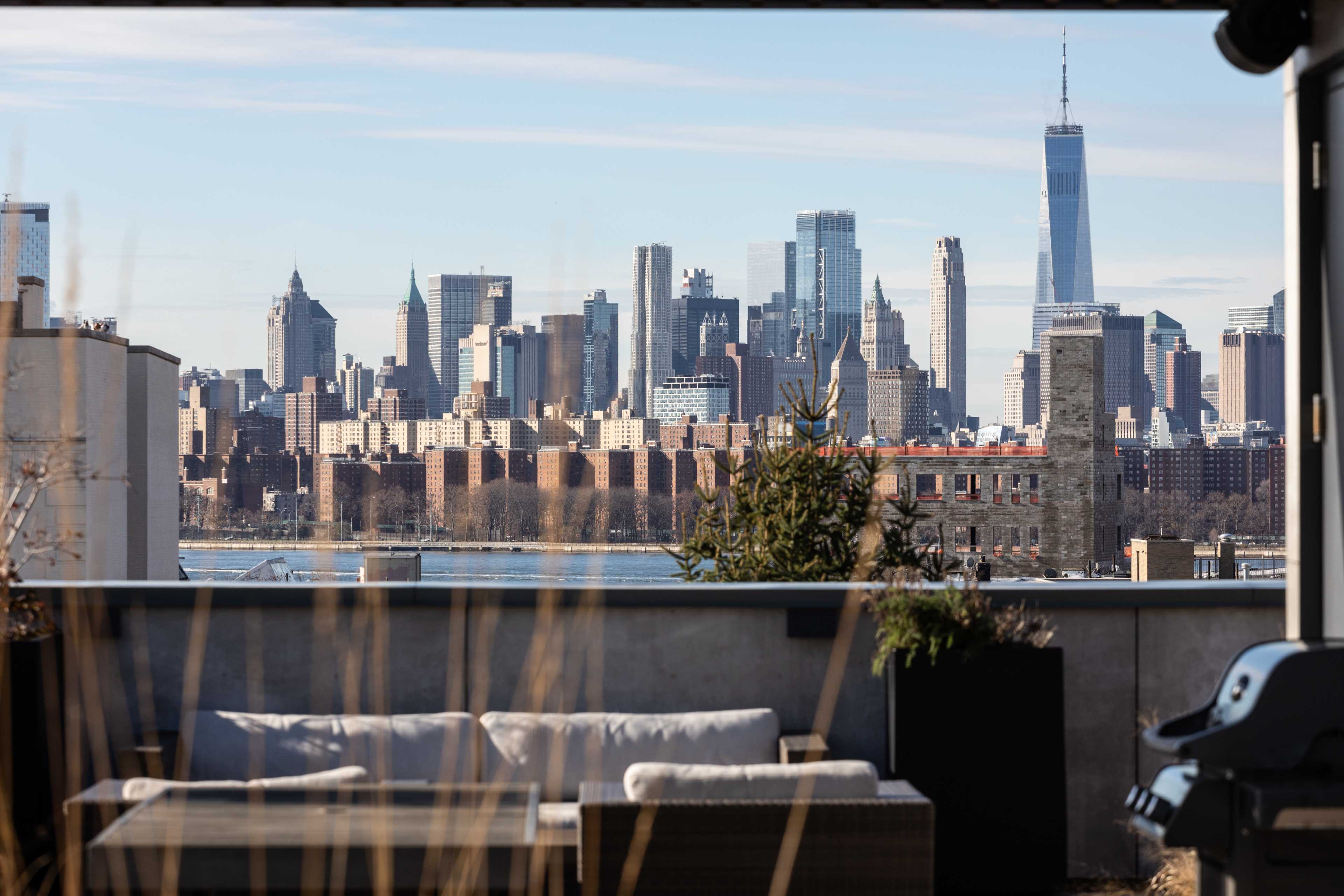 A view of the Manhattan skyline is framed by a rooftop terrace with a couch and potted plants.