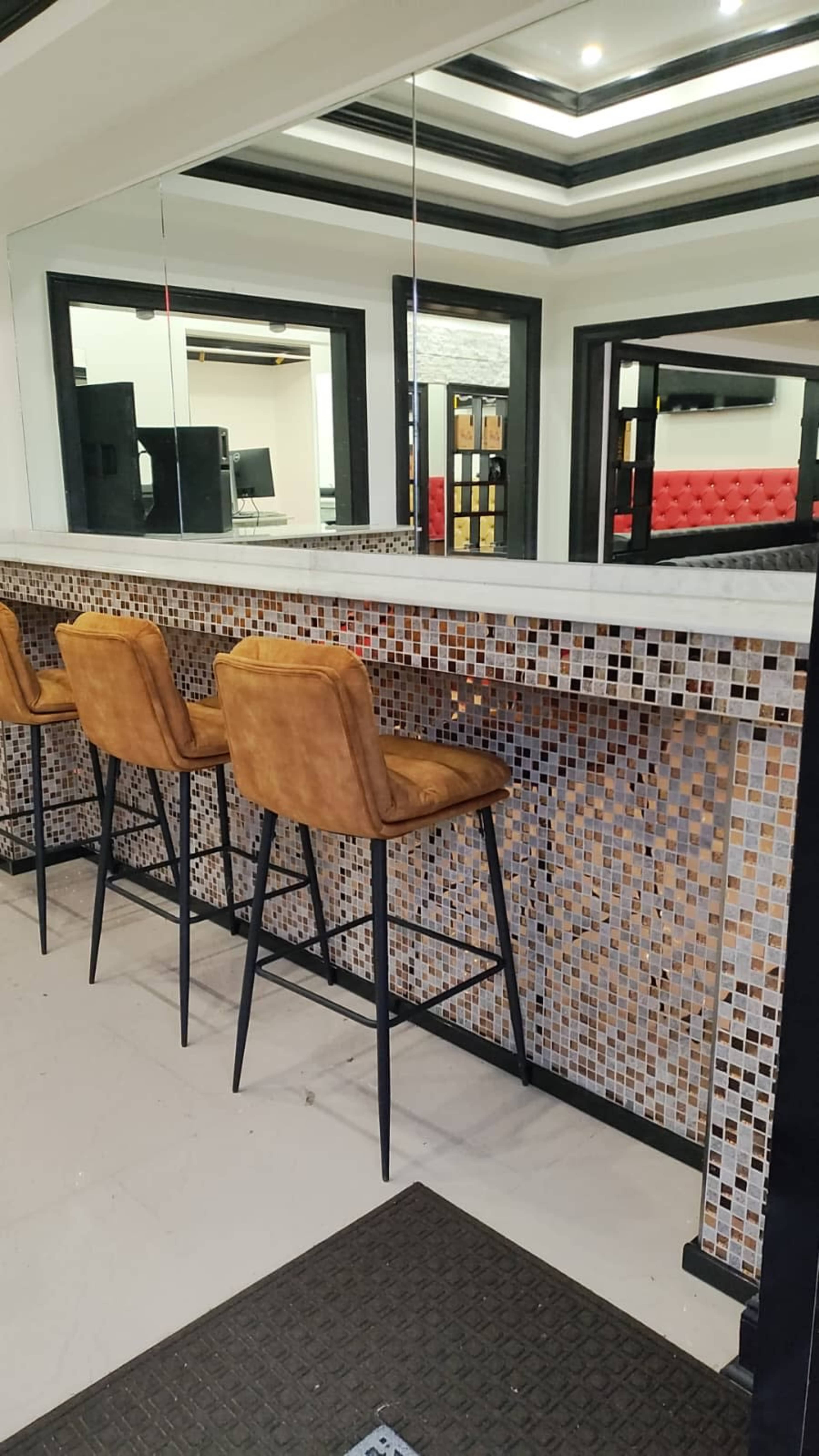 The image shows a modern bar area with three tall, upholstered stools arranged in front of a mosaic-tiled wall and a mirrored surface above.