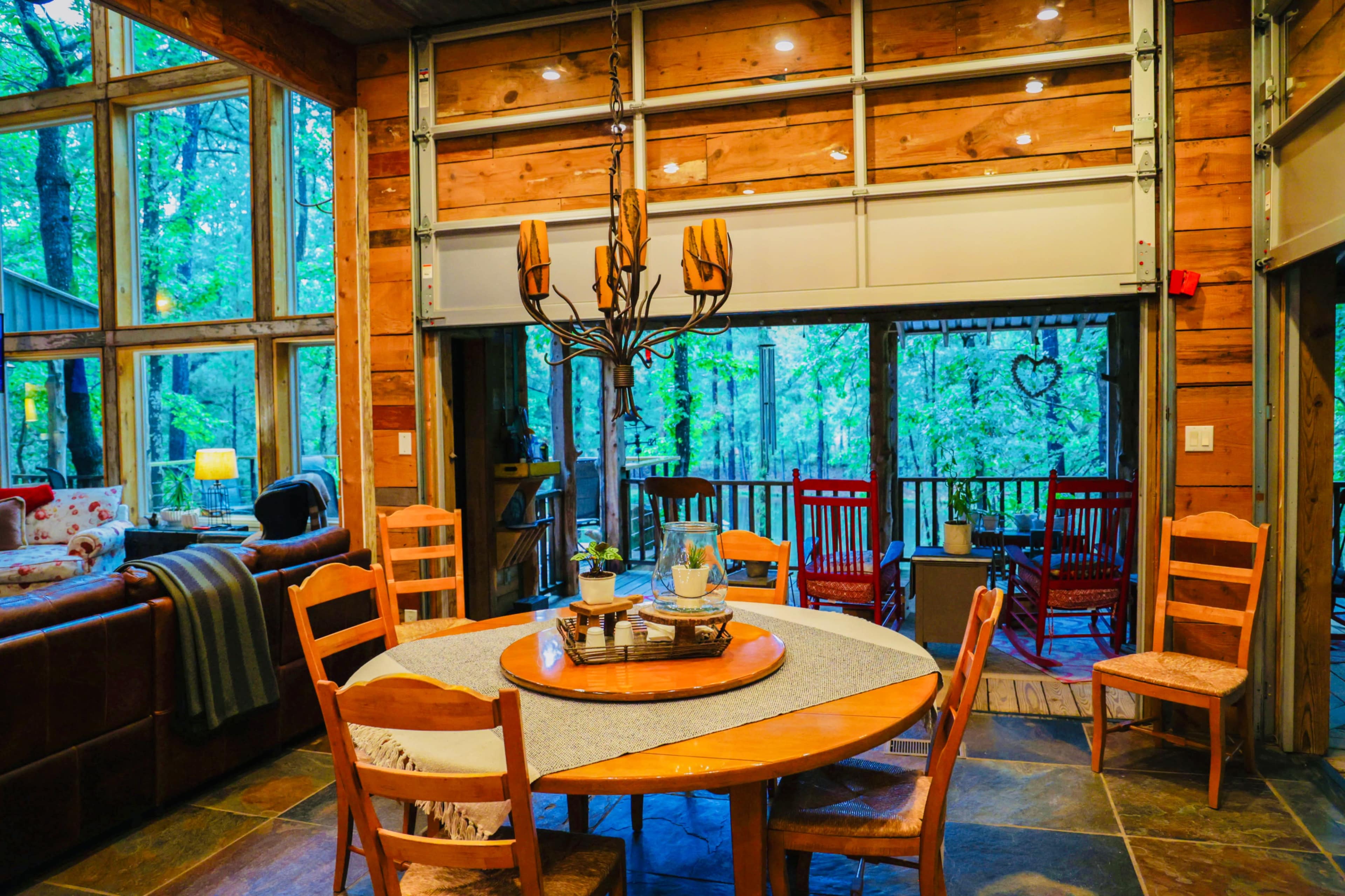 A round dining table with wooden chairs is situated in a cabin-like interior that features large windows overlooking a forested area.