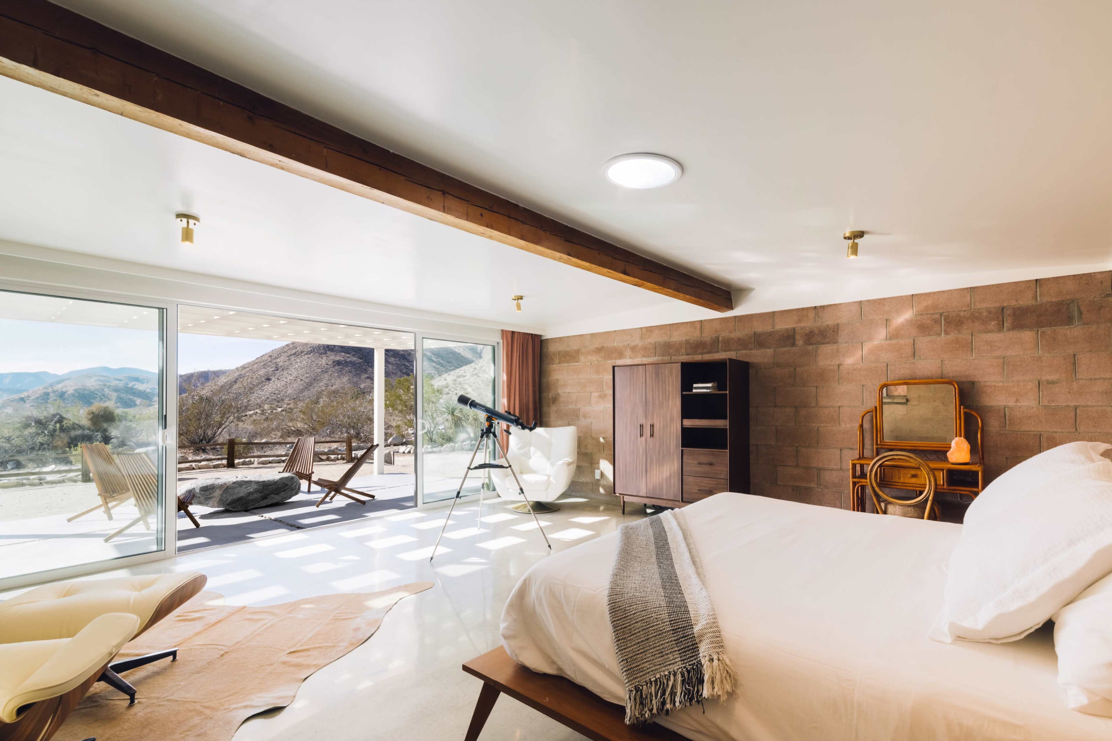 The image depicts a modern bedroom with a large window showcasing a mountainous landscape, featuring a telescope and minimalist furniture.