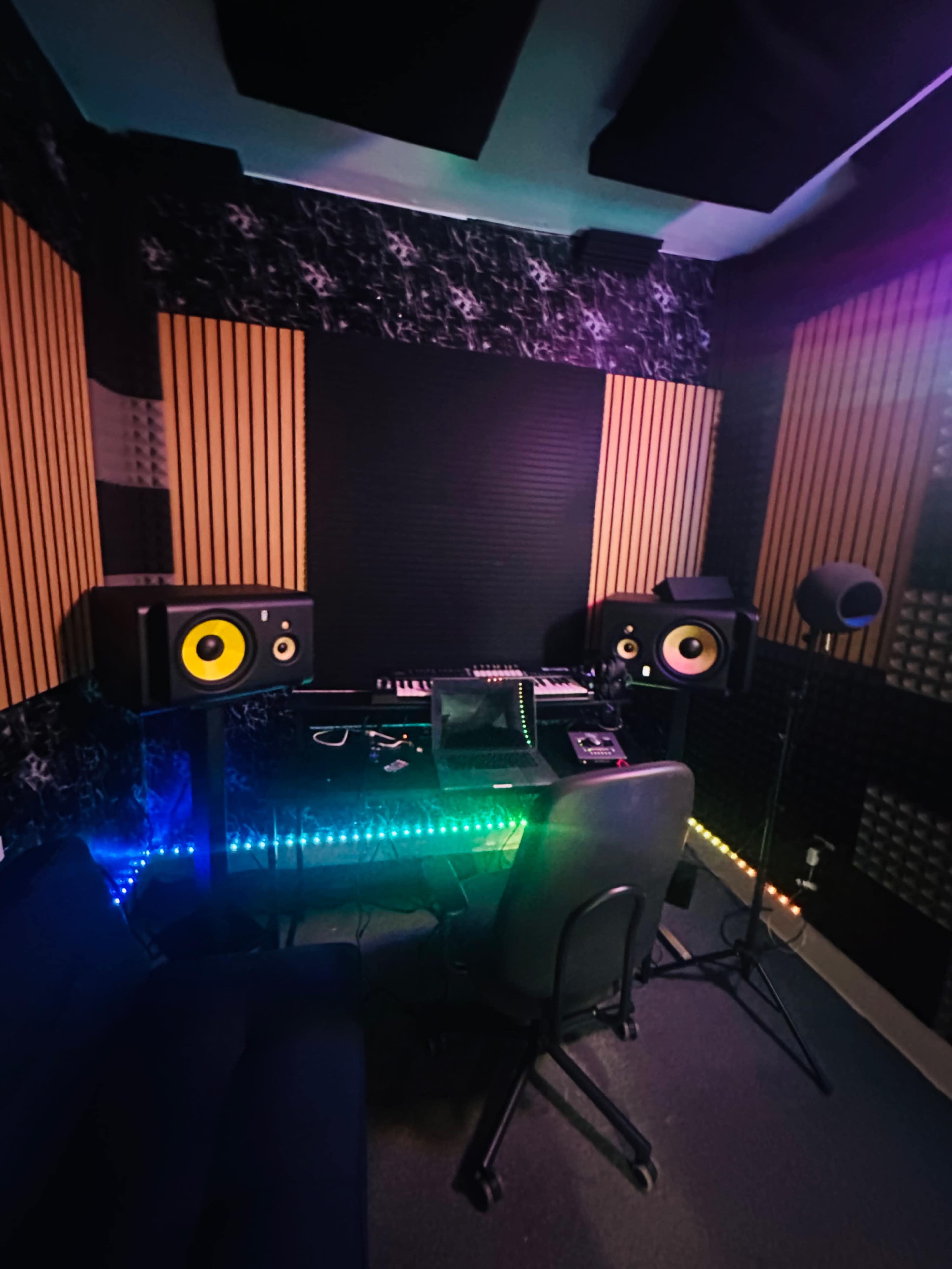 The image shows a music studio featuring a desk with audio equipment, two speaker monitors, and colorful LED lights along the floor.