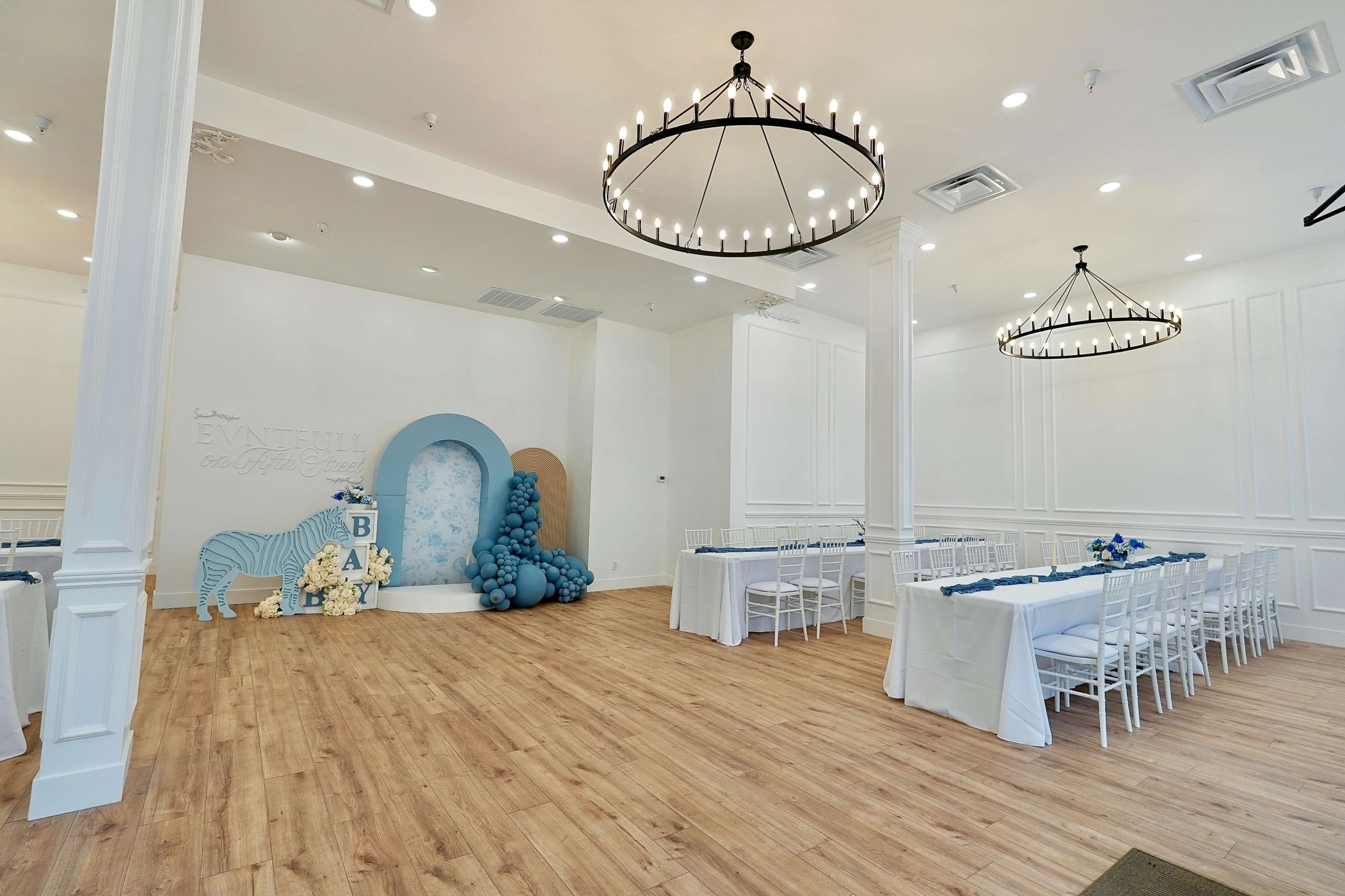 A spacious event room features white walls, wooden flooring, and a decorative backdrop with blue balloons and floral arrangements.
