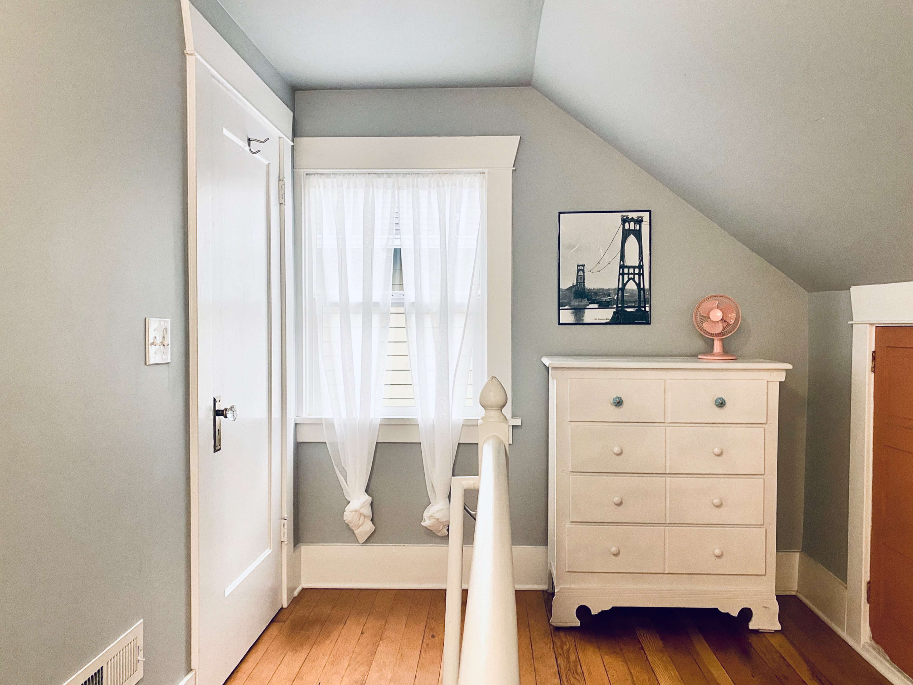 A corner of a room featuring a white dresser, a framed picture of a bridge on the wall, sheer curtains over a window, and a small pink fan on top of the dresser.