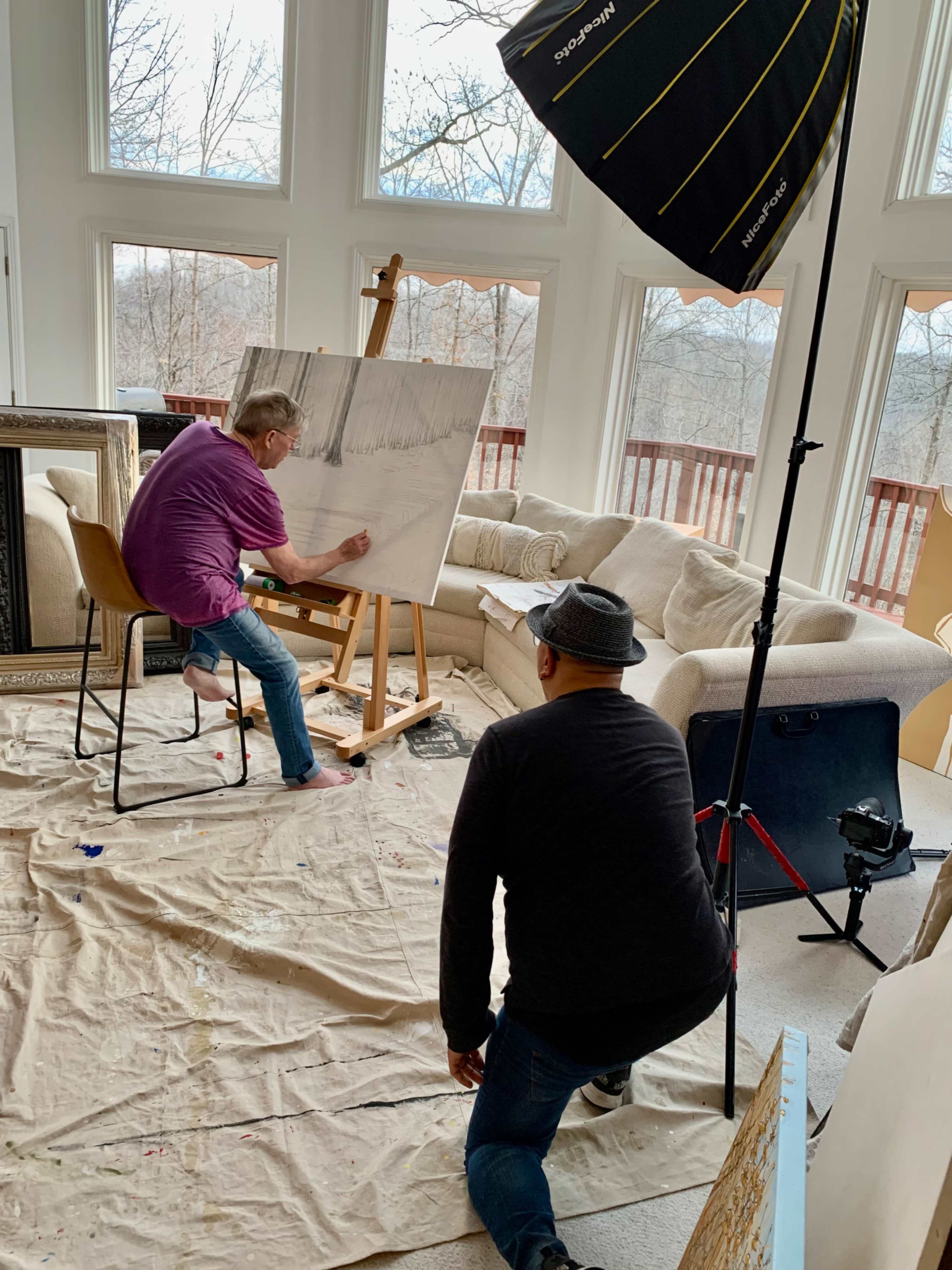 An artist is painting on an easel in a bright room while a person observes from the floor, both surrounded by art supplies and furniture.