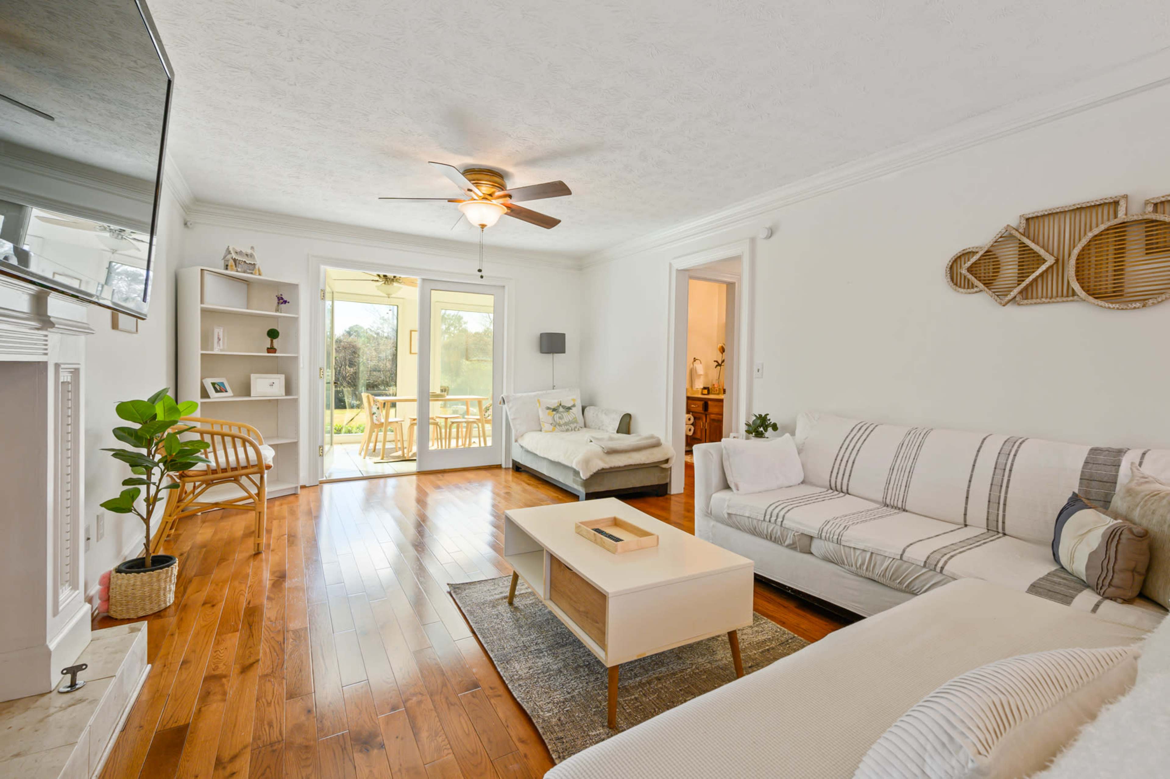 A bright living room with a wooden floor, a ceiling fan, a white sectional sofa, and a coffee table, while large doors lead to a patio area.