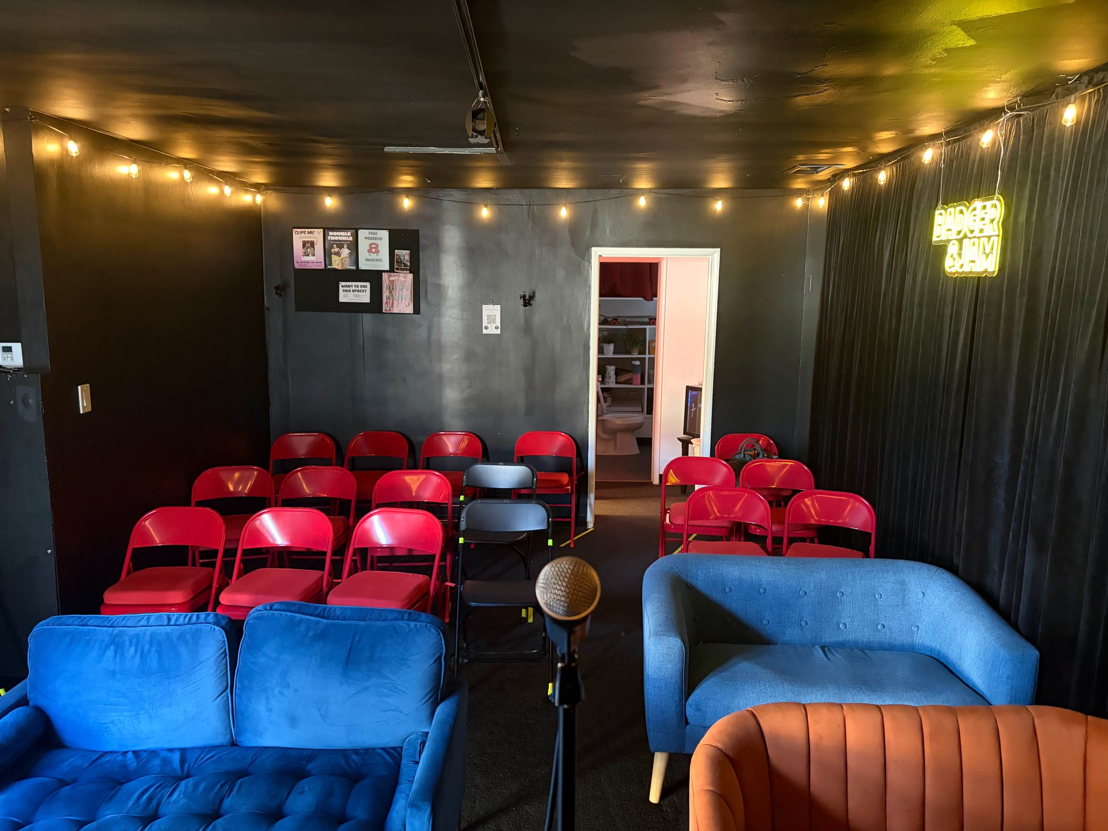 The image shows a small, dimly-lit performance space featuring rows of red chairs facing a microphone and blue couches, with string lights illuminating the walls.