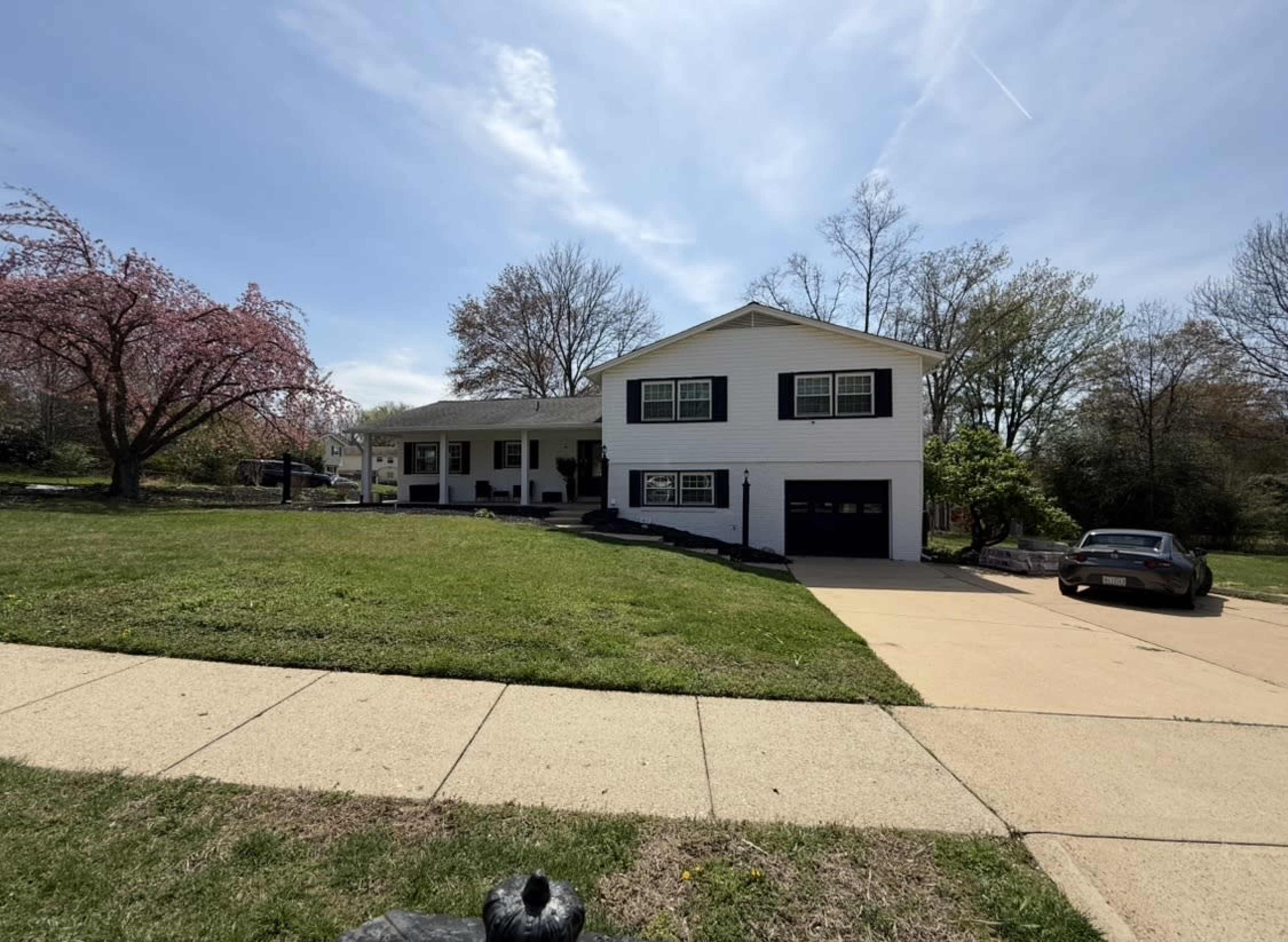 A two-story white house with a black garage door is situated on a lawn surrounded by trees and a blooming pink cherry blossom.