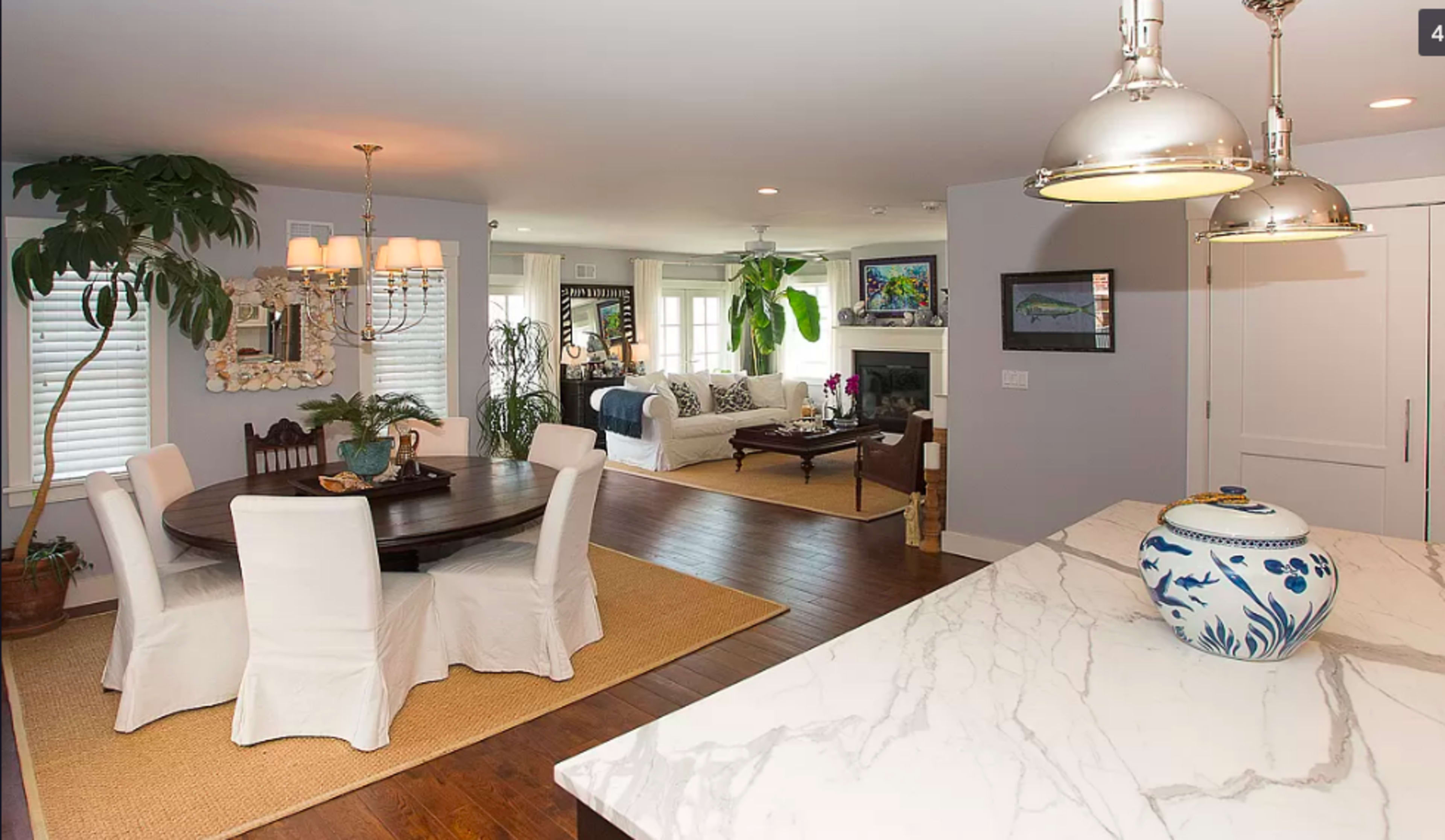 The image shows a bright, open-concept dining area with a round table and white chairs, adjacent to a living space featuring a sofa, plants, and a television.