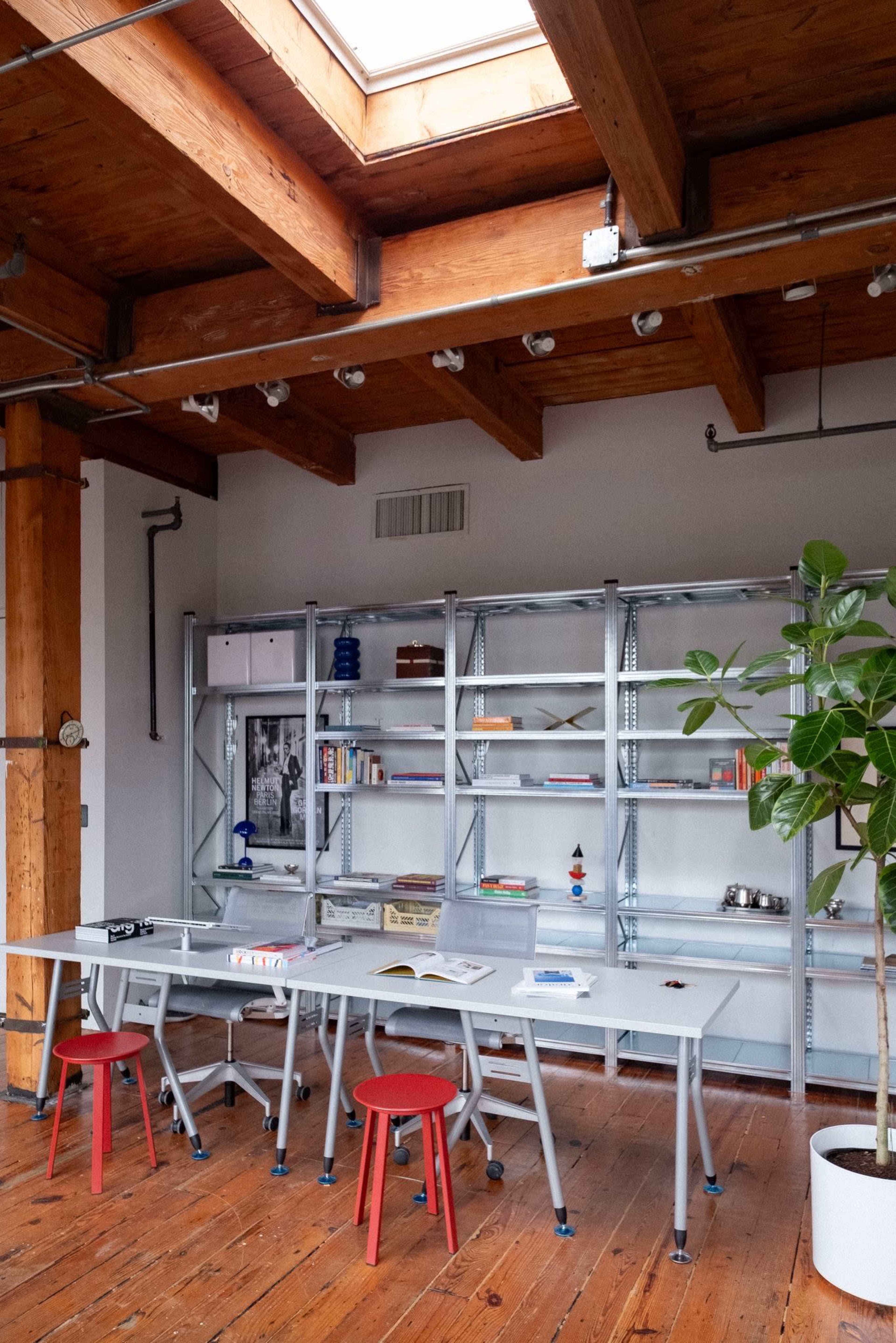 The image shows a modern workspace featuring a light-colored table, red stools, a metal bookshelf filled with books and decorative items, and large wooden beams in the ceiling.