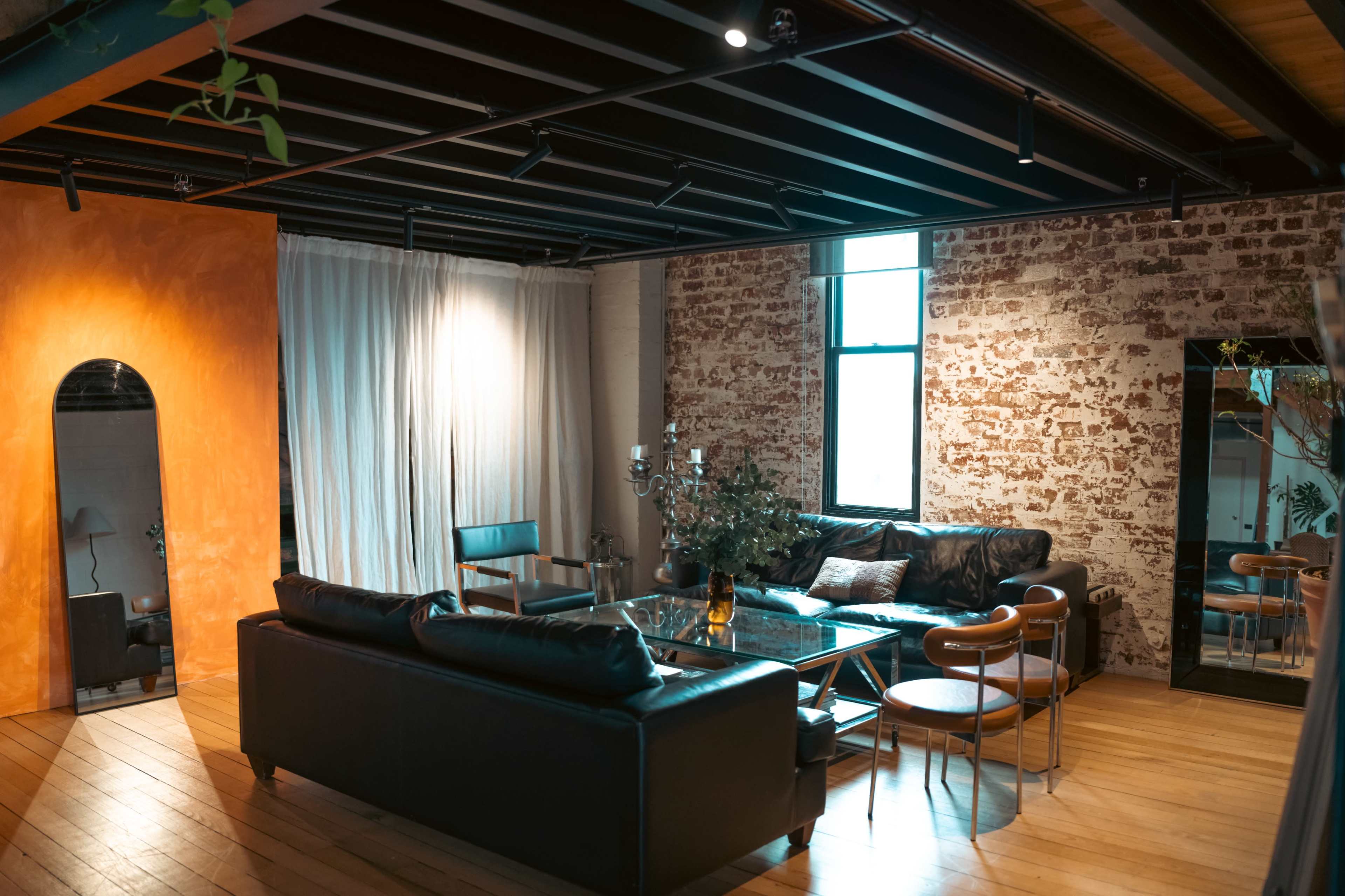 The image shows a modern living room with a mix of exposed brick and orange-painted walls, featuring a black leather sofa, a glass coffee table, and various decorative elements.