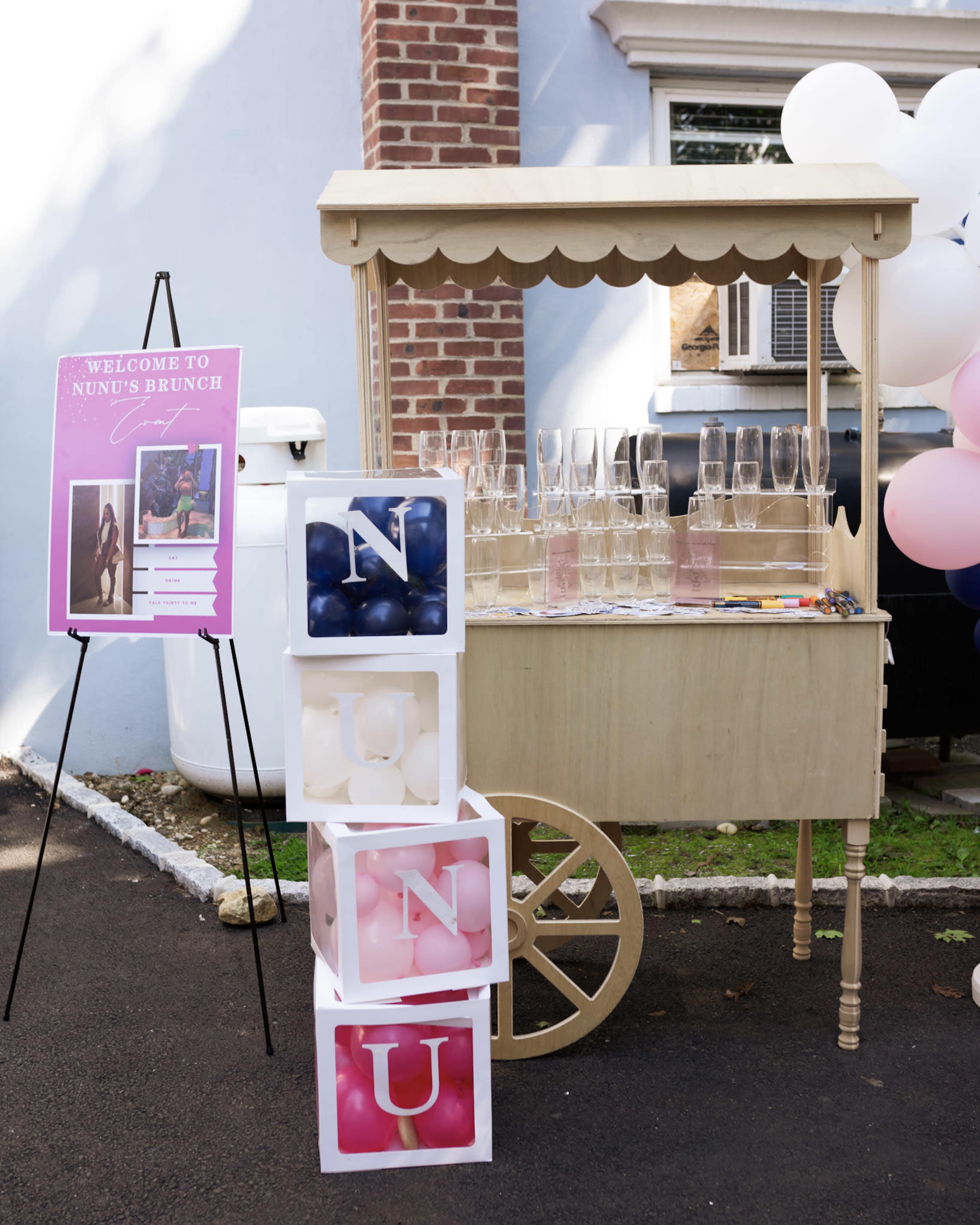 A decorated beverage cart with glasses and a sign welcoming guests to "Nunu's Brunch" is displayed alongside stacked blocks featuring the letters "N," "U," "N," and "U."