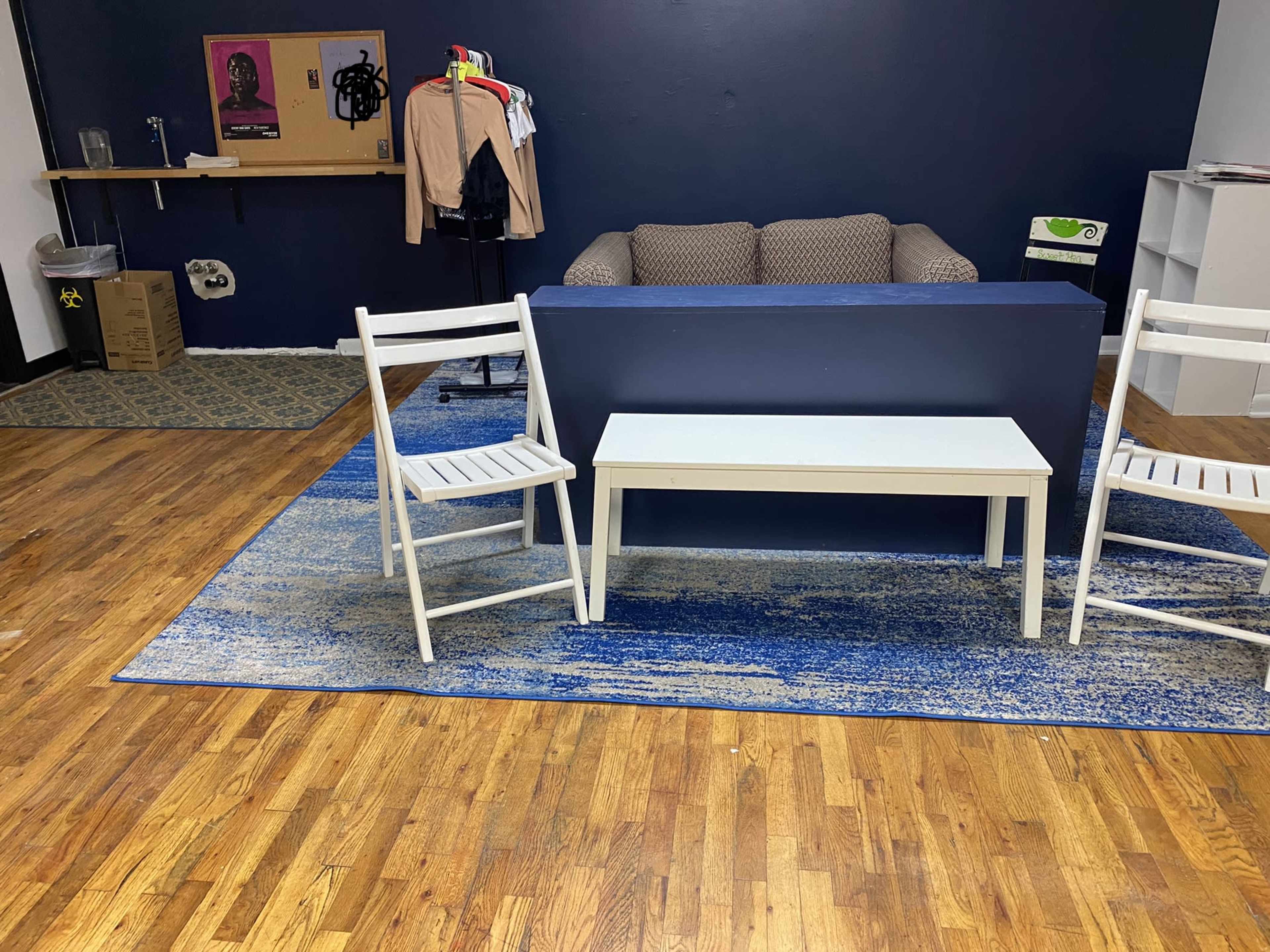 A room with a brown couch against a blue wall, two white chairs, and a light-colored table on a blue rug.