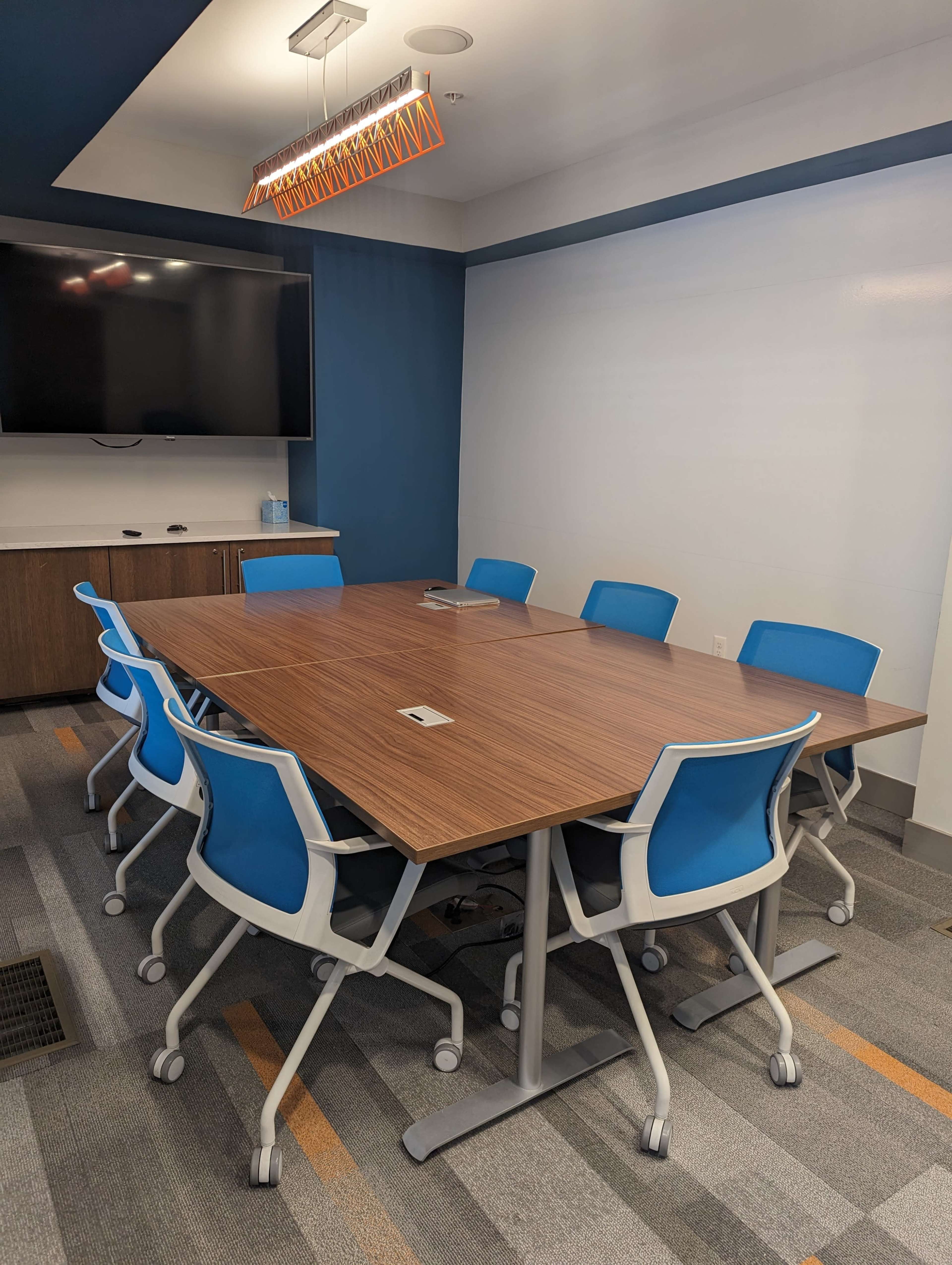A modern conference room features a large brown table surrounded by eight blue and white rolling chairs, with a wall-mounted screen in the background.