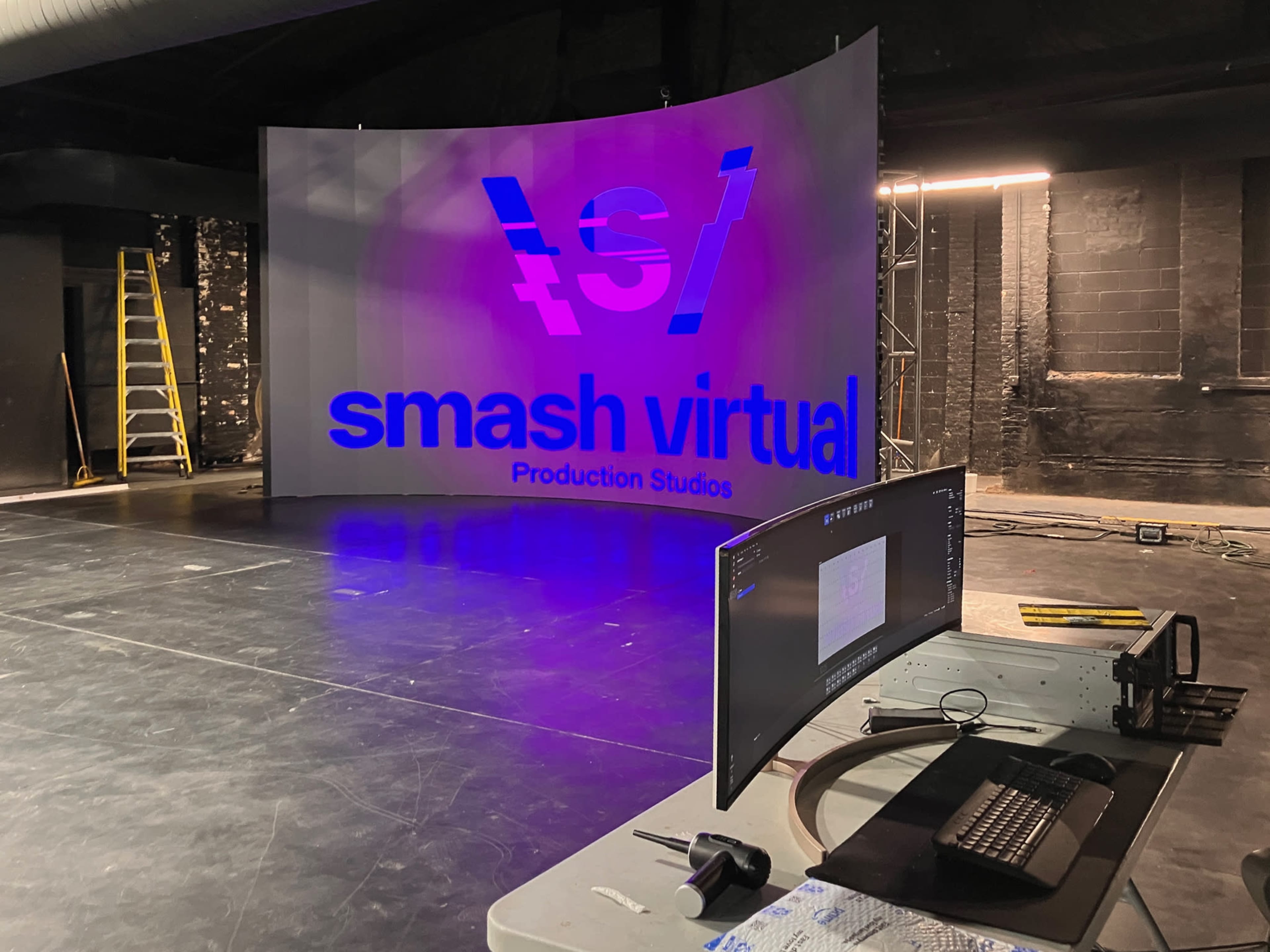 The image shows a production studio featuring a large backdrop with the text "smash virtual" illuminated in blue and white, along with a computer setup in the foreground.