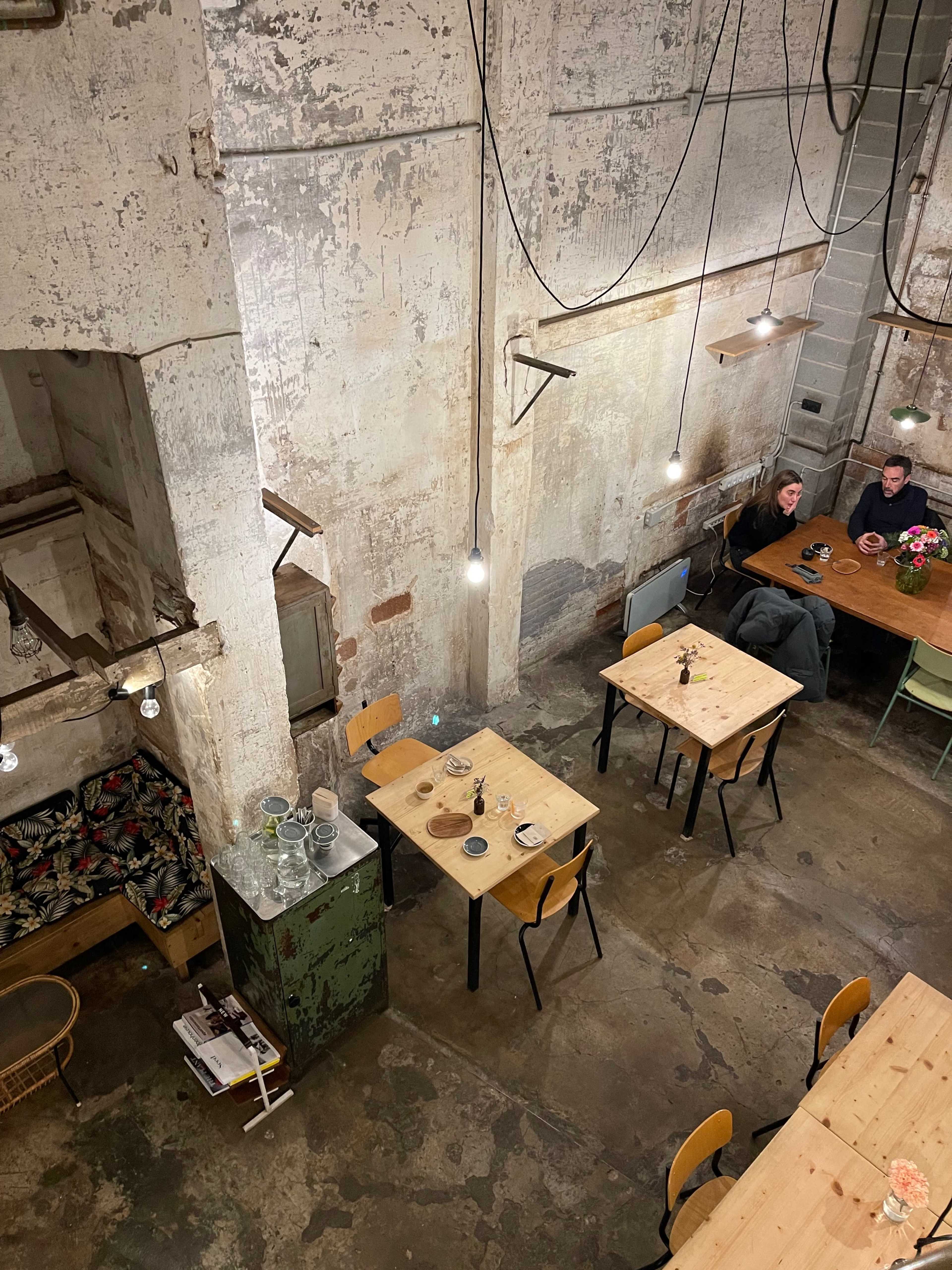 A dimly lit café features wooden tables, a vintage green cabinet, and patrons seated in a rustic setting with exposed walls and hanging light fixtures.