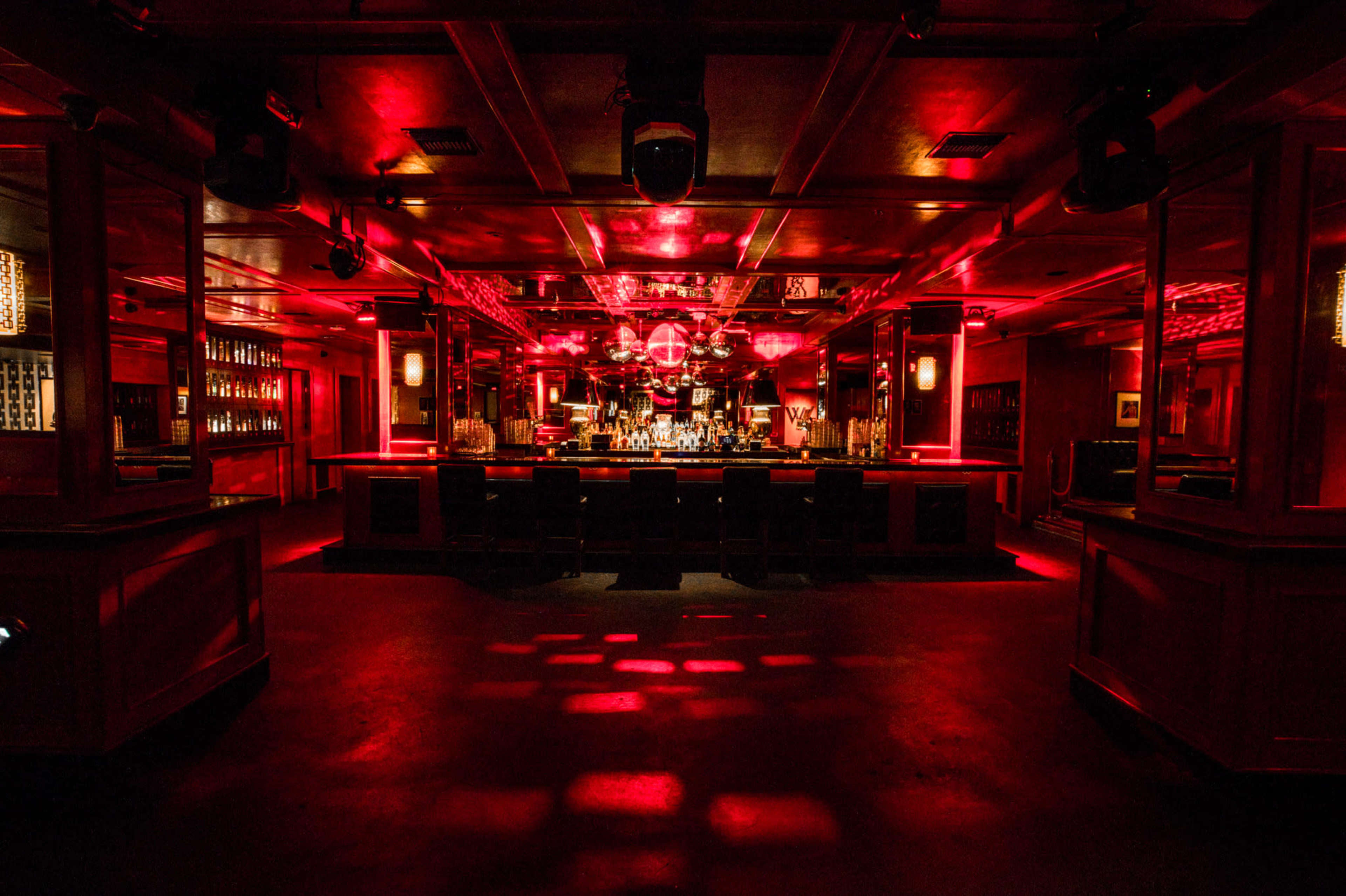 The image shows a dimly lit bar with red lighting, a central mirrored disco ball, and a long bar counter lined with stools.