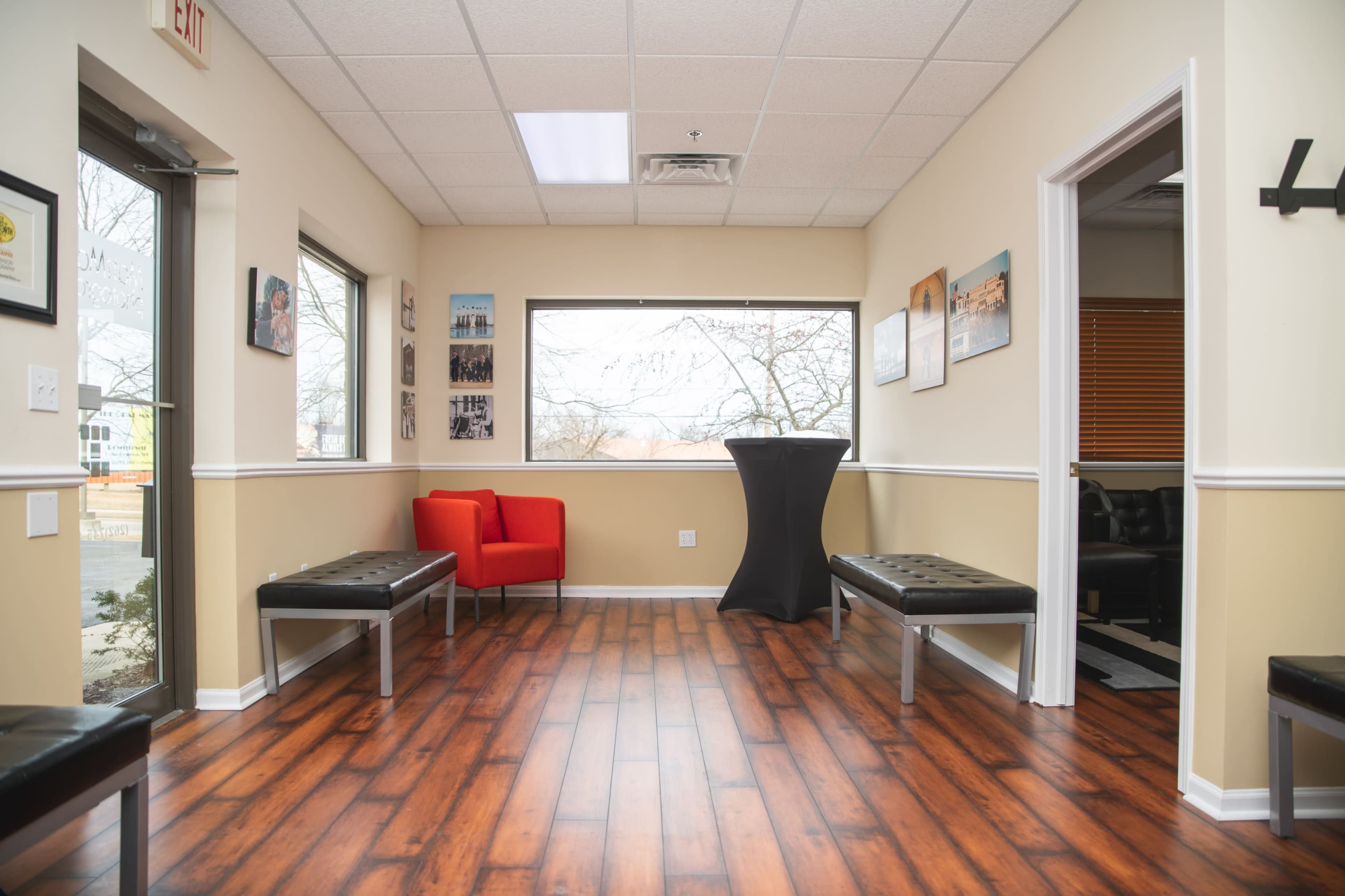 A waiting room with wooden flooring, two benches, a red chair, and a tall black table near a large window.