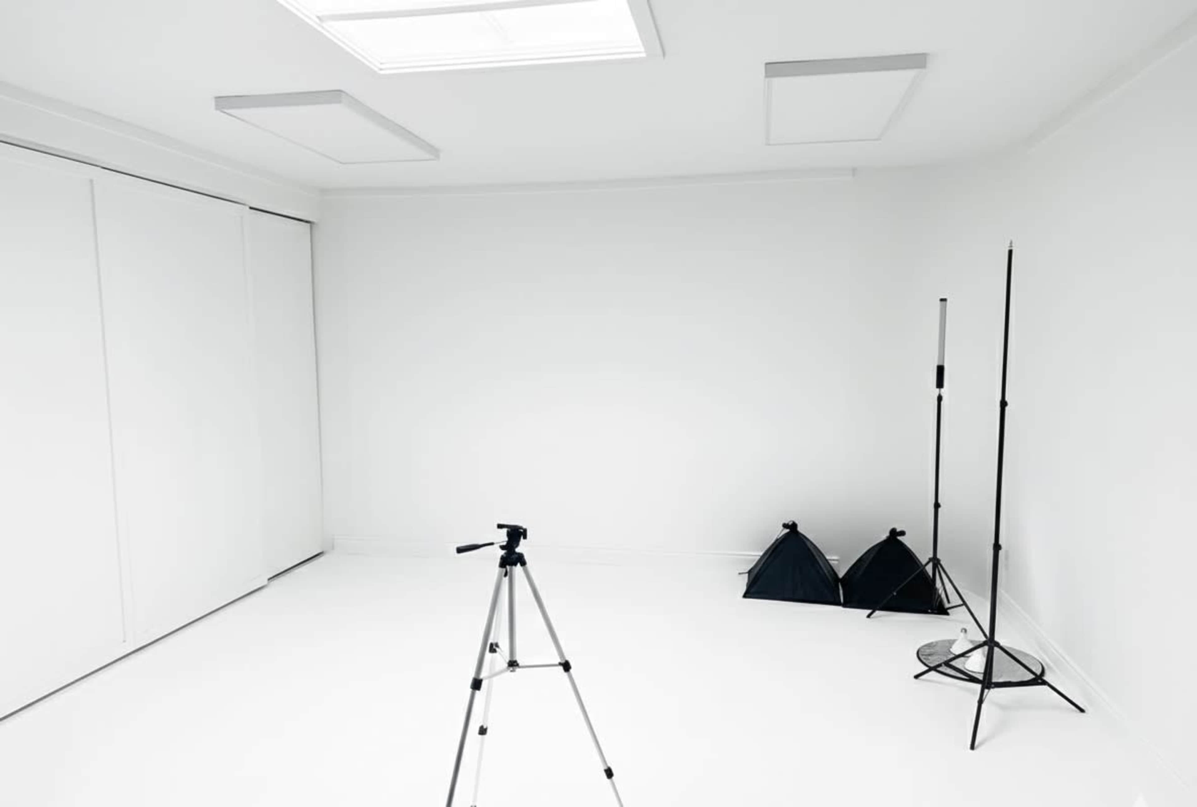 The image shows a minimalistic white studio space with a camera tripod in the center and two light stands in the corner.