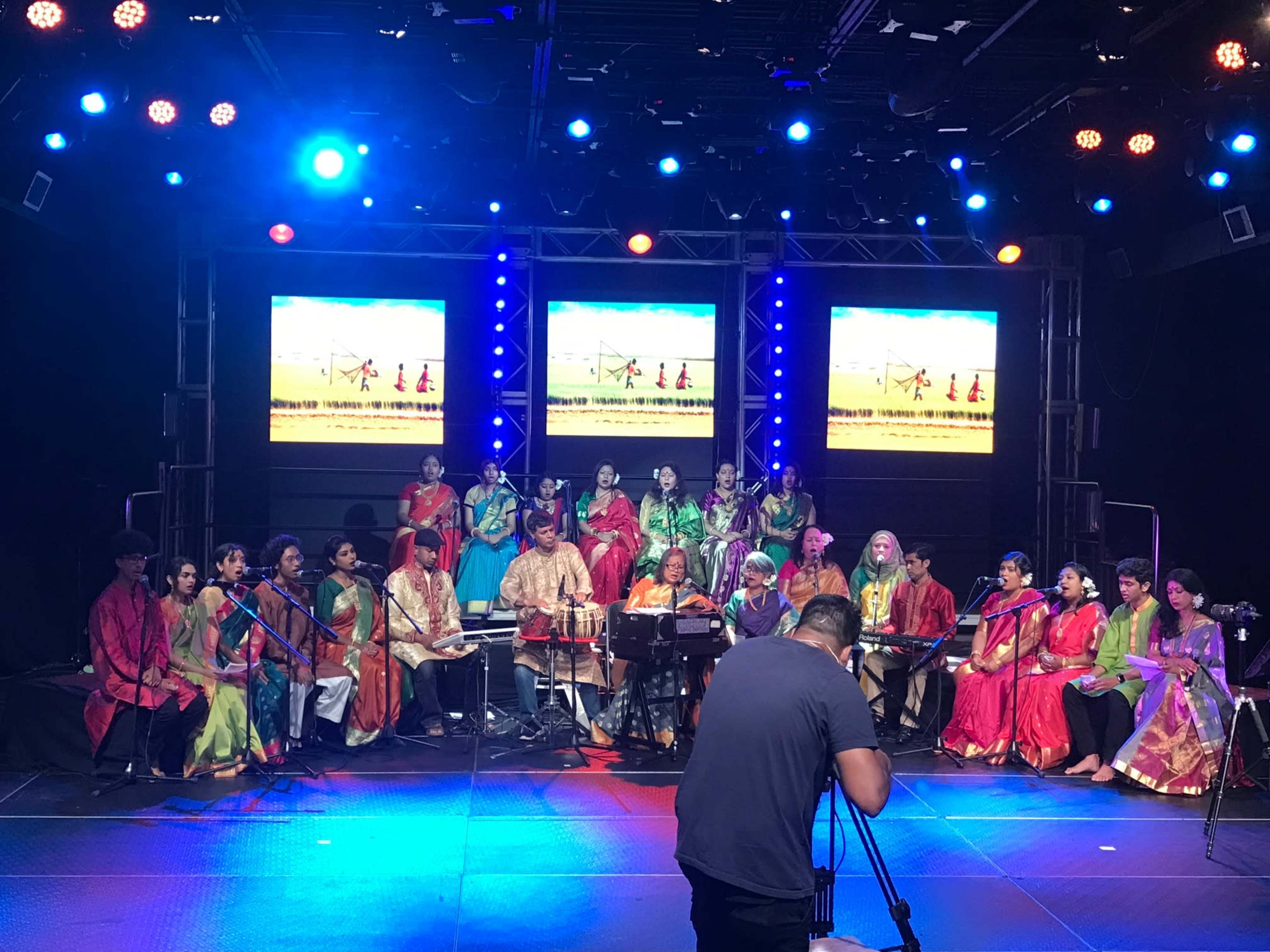 A diverse group of musicians and singers dressed in colorful traditional attire performs on stage with instruments and lighting.