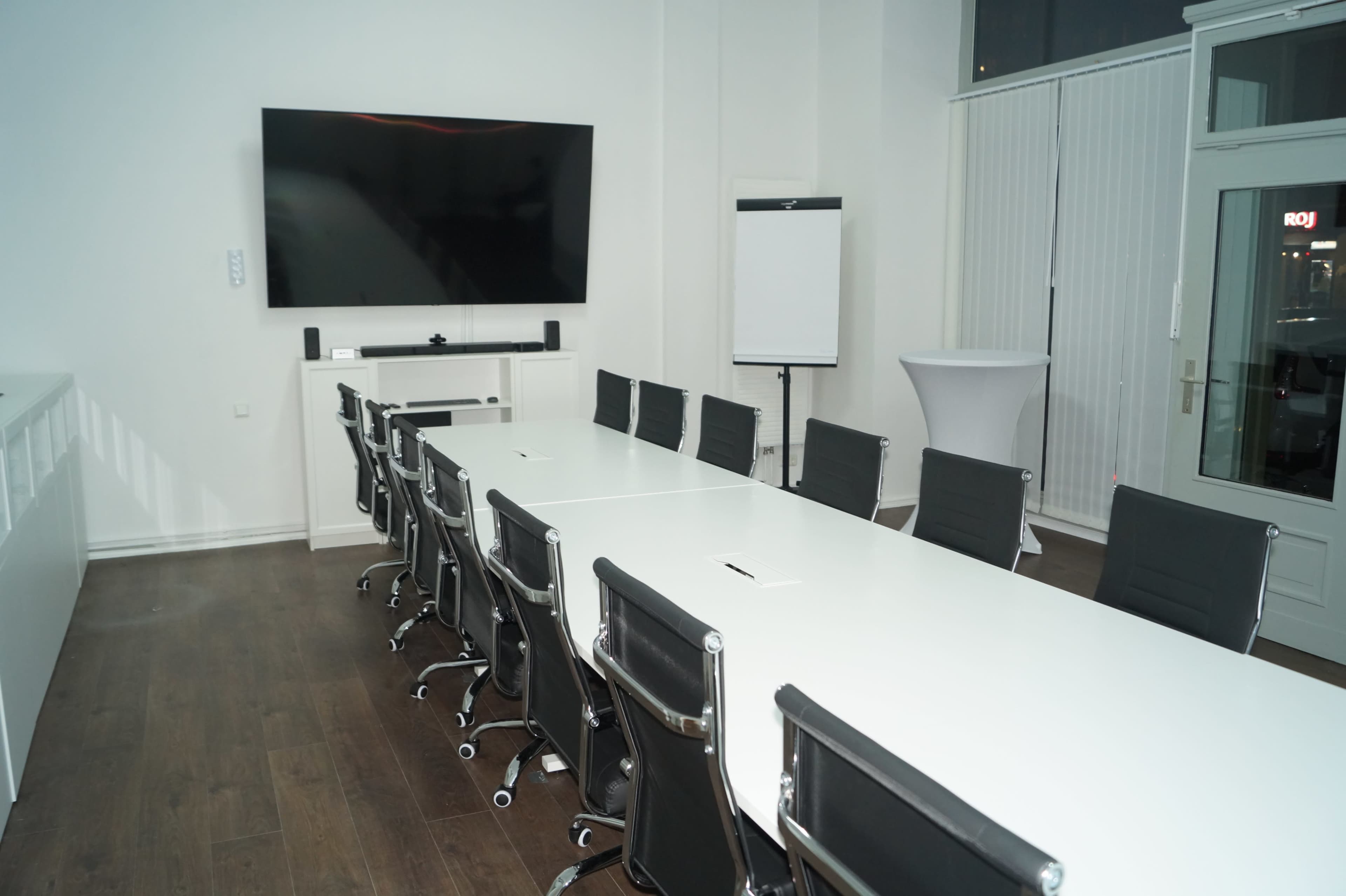 A modern conference room features a long white table with black rolling chairs and a large screen mounted on the wall.