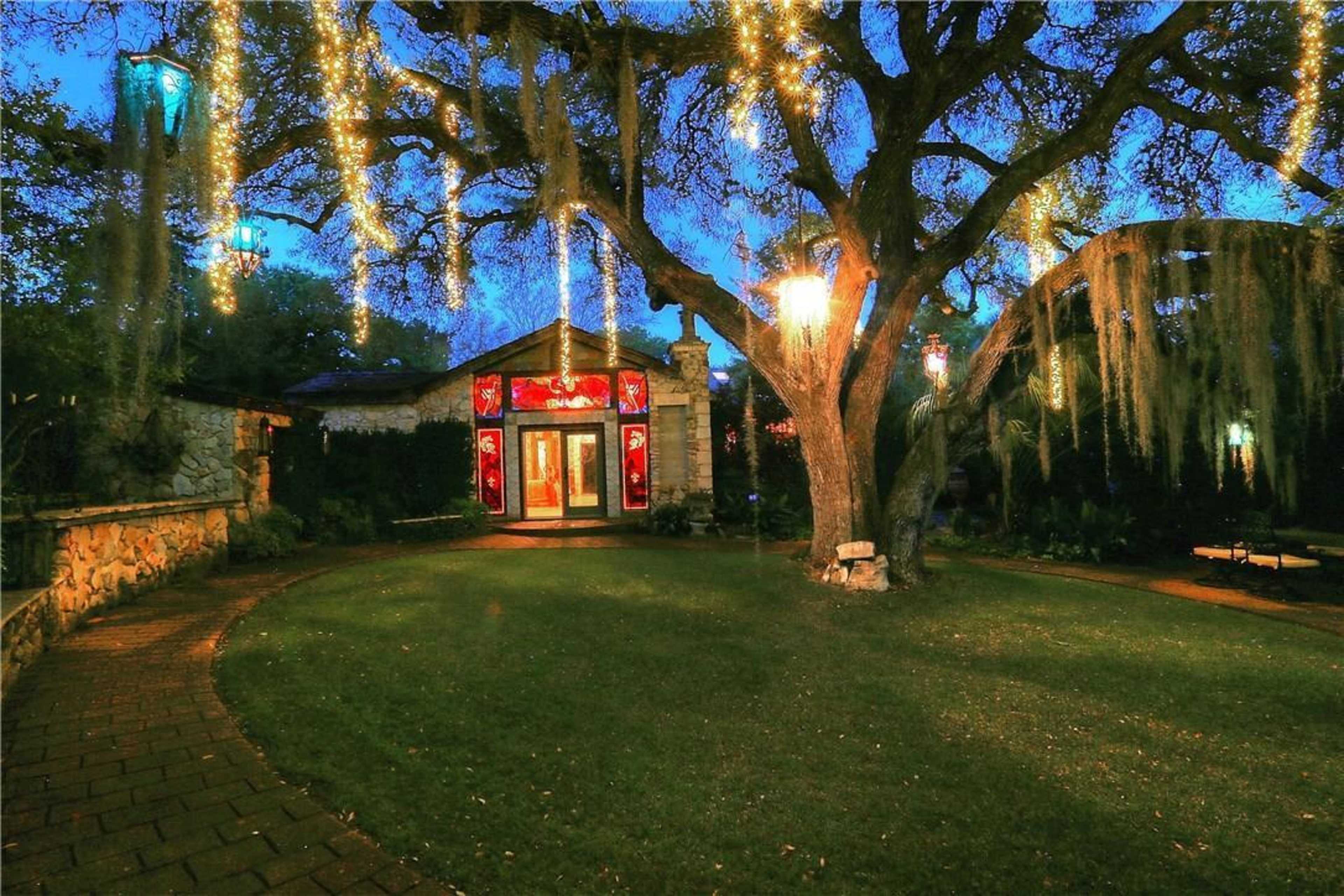 A large tree draped with Spanish moss and string lights stands next to a stone path leading to a building with red windows in a landscaped garden at twilight.