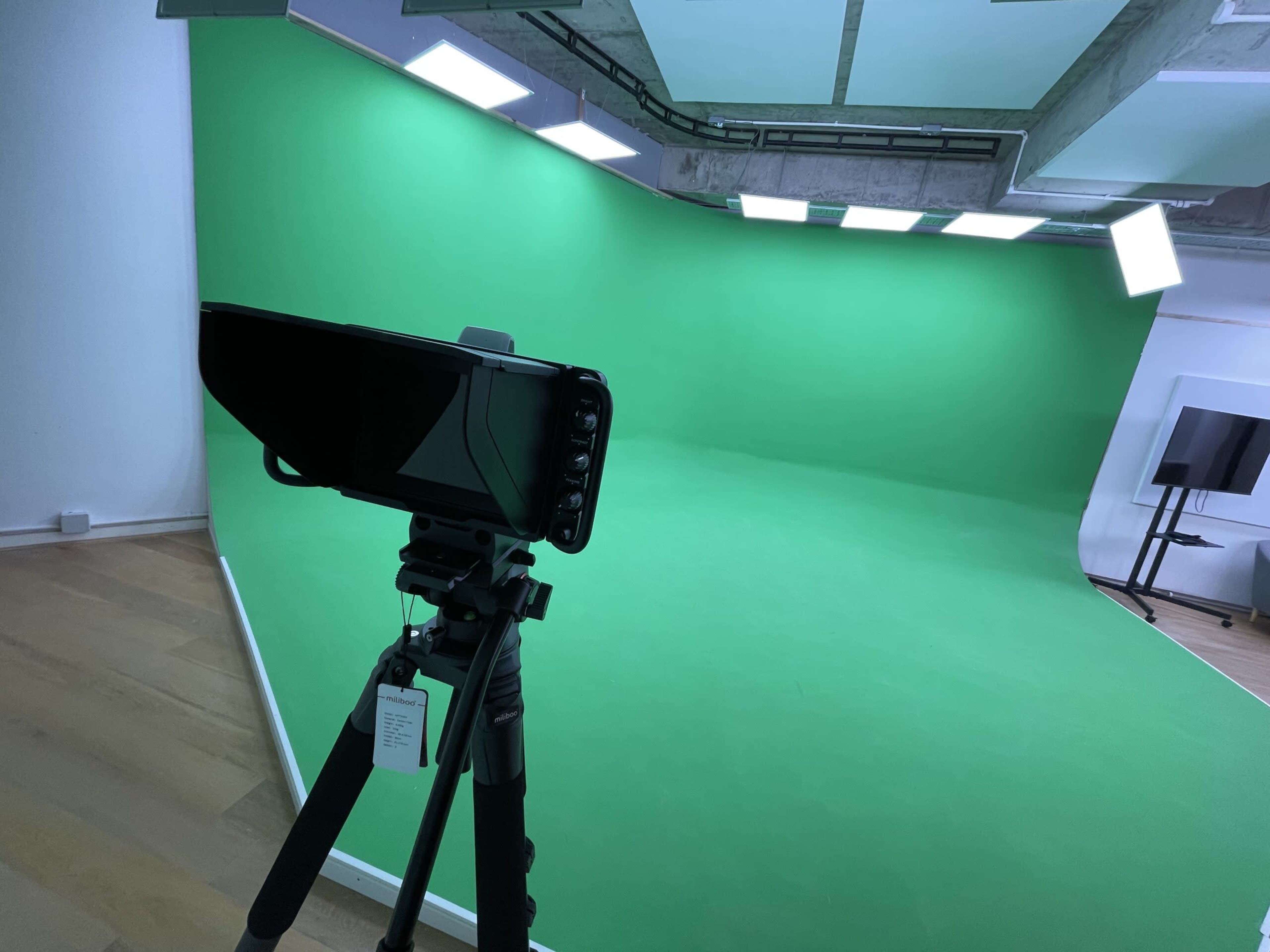 The image shows a green screen setup with a camera mounted on a tripod in a studio environment.