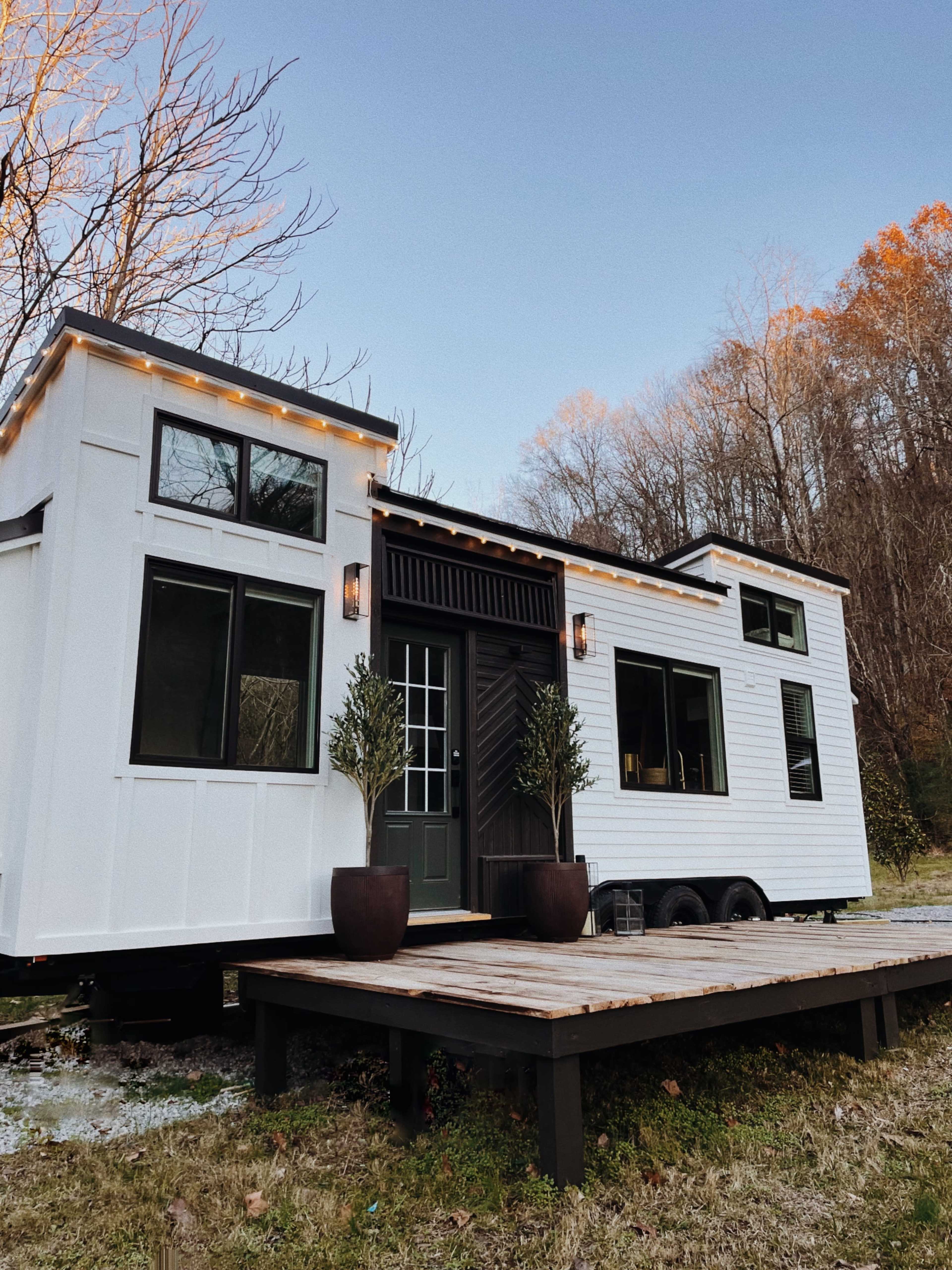 A modern tiny house on wheels with a wood deck, surrounded by trees and a clear sky.