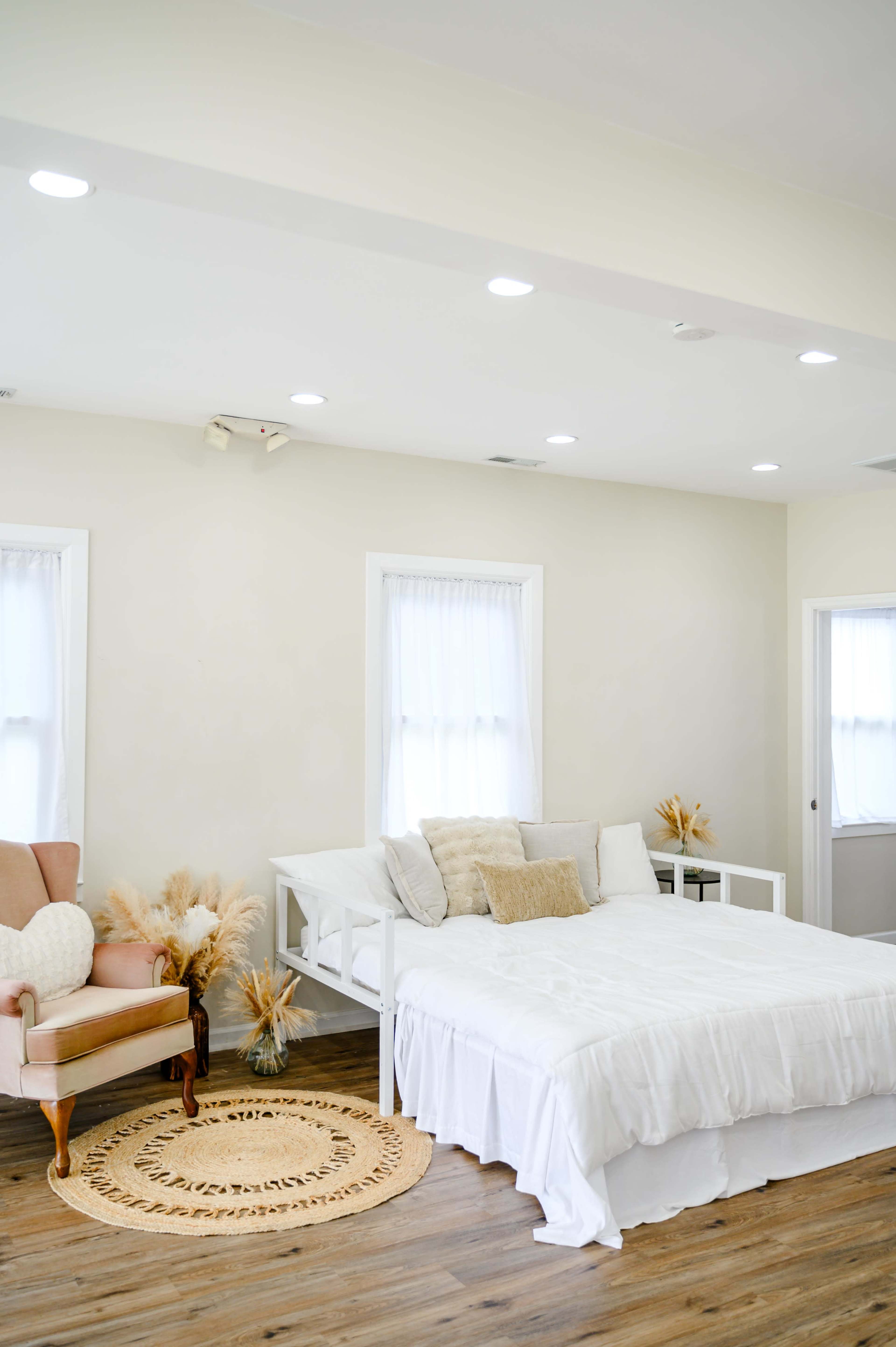 The image shows a bright, minimalist bedroom with a white bedspread, a pink armchair, and decorative plants arranged on the floor.
