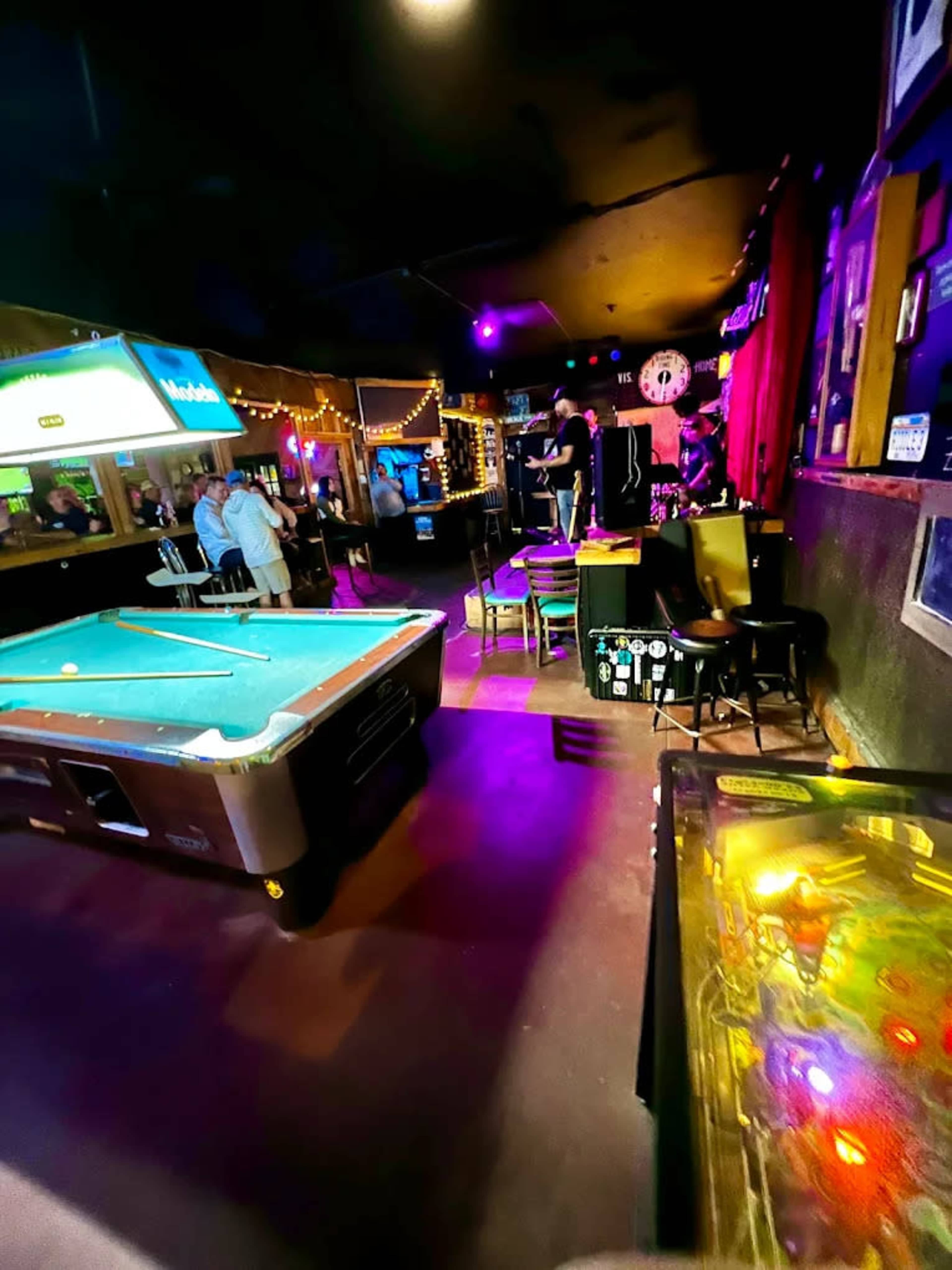 The image shows a dimly lit bar setup featuring a pool table, a pinball machine, and patrons socializing in the background.