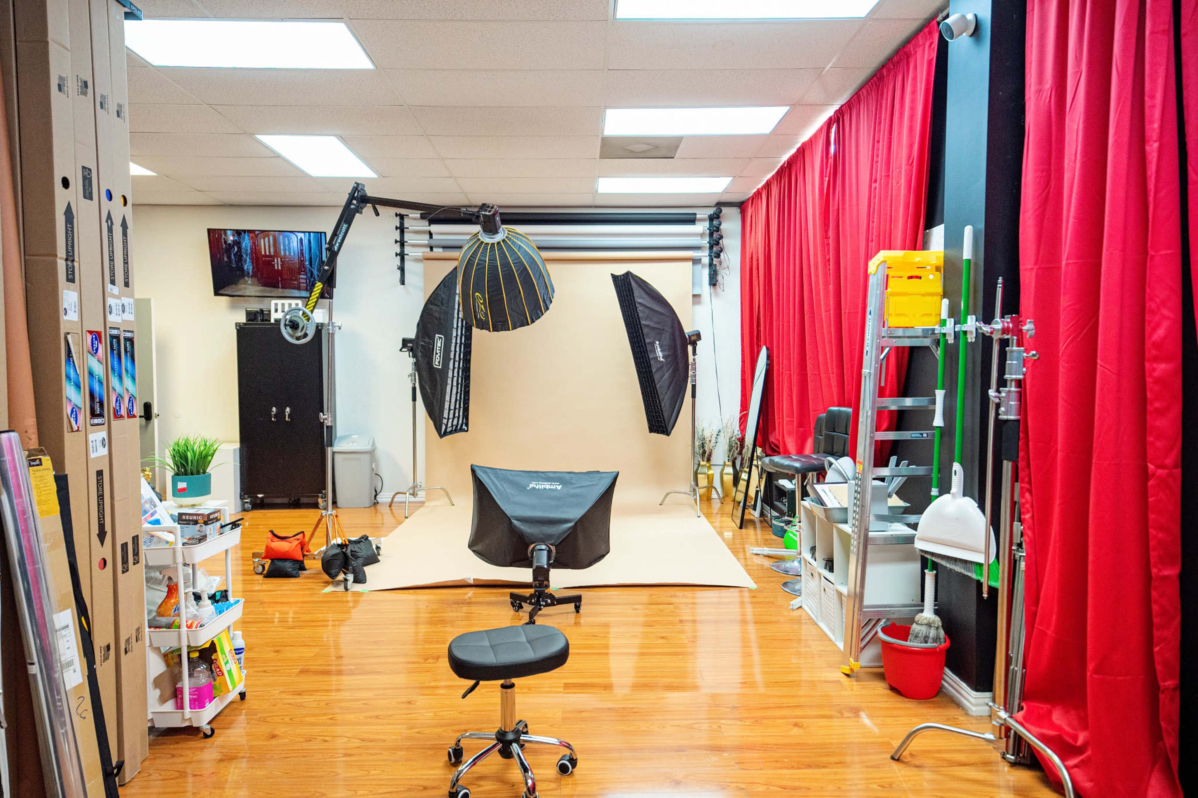 The image shows a photography studio with a beige backdrop, various lighting equipment, a chair, and shelves filled with supplies, framed by red curtains.