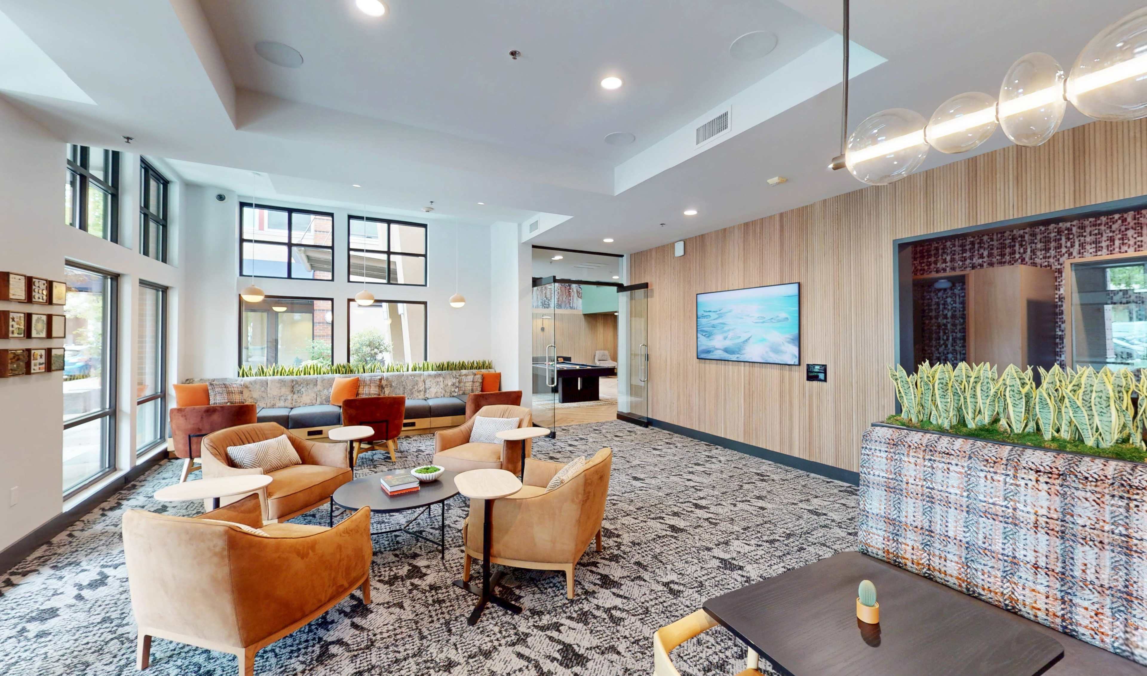 The image shows a modern lounge area with comfortable seating, large windows, and a central coffee table.