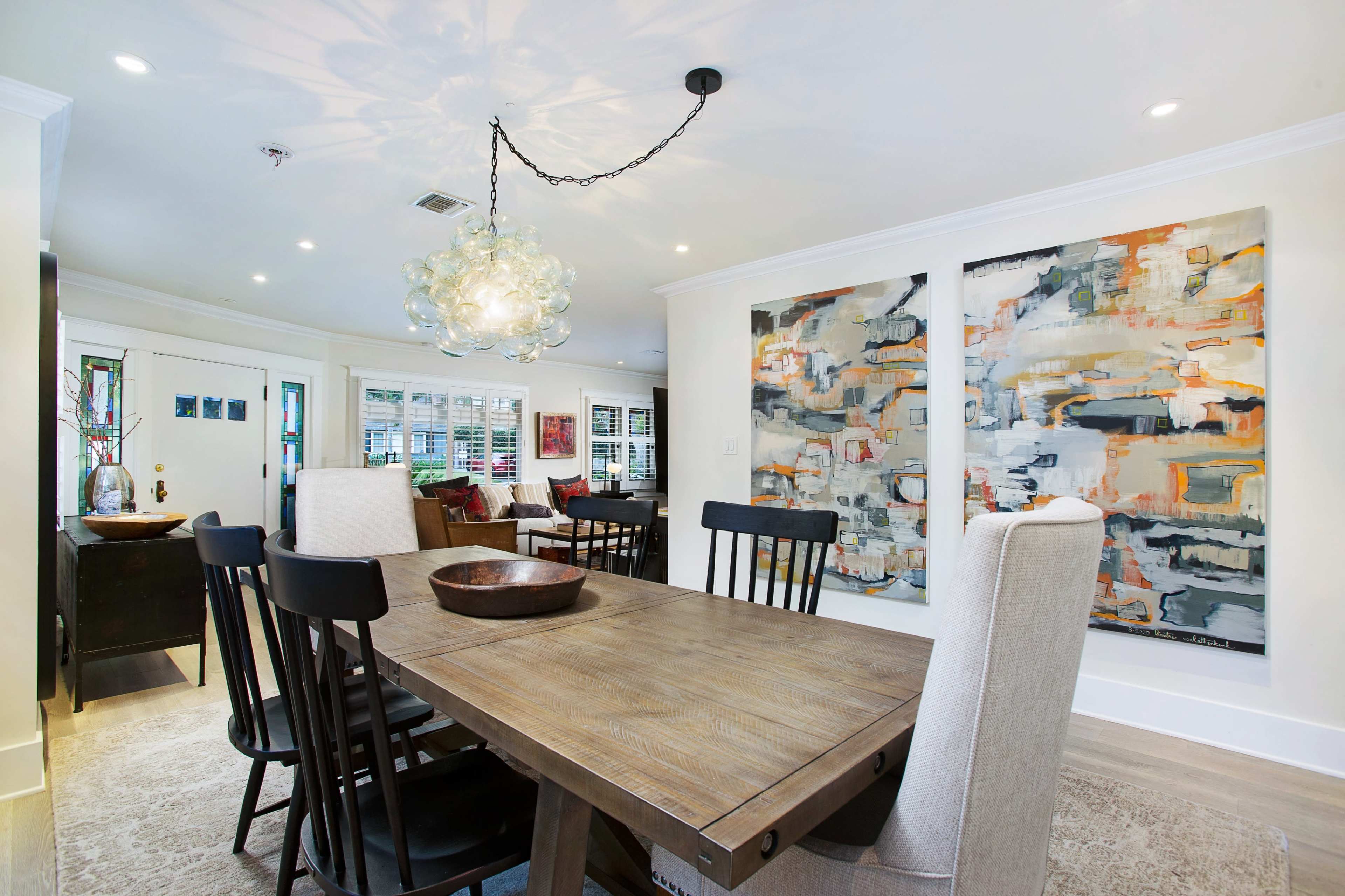 A modern dining area features a wooden table surrounded by black and beige chairs, illuminated by a unique chandelier, with two large abstract paintings hanging on the wall.